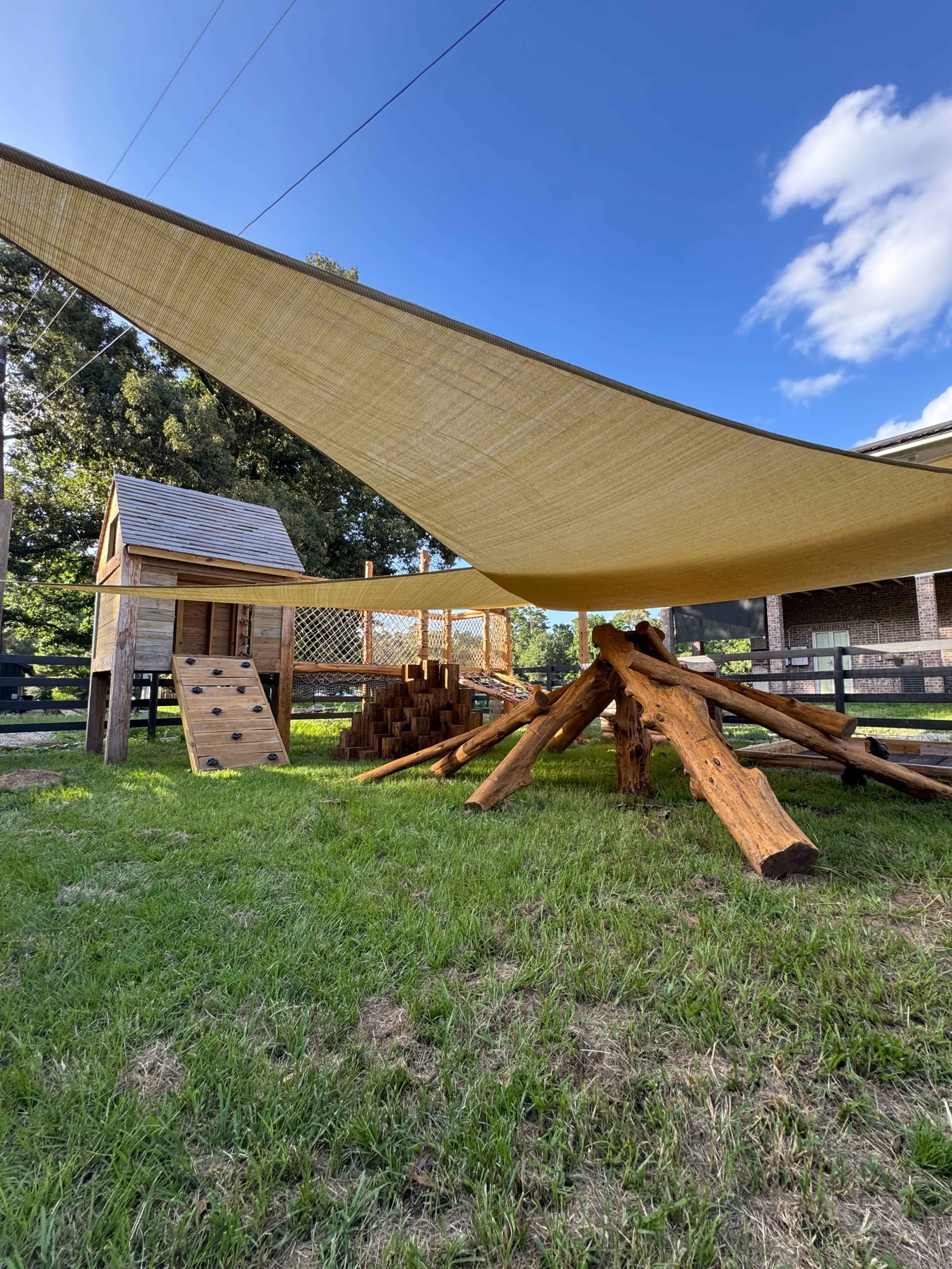 A large shade structure made of fabric is supported by logs above a wooden play area in a grassy field.