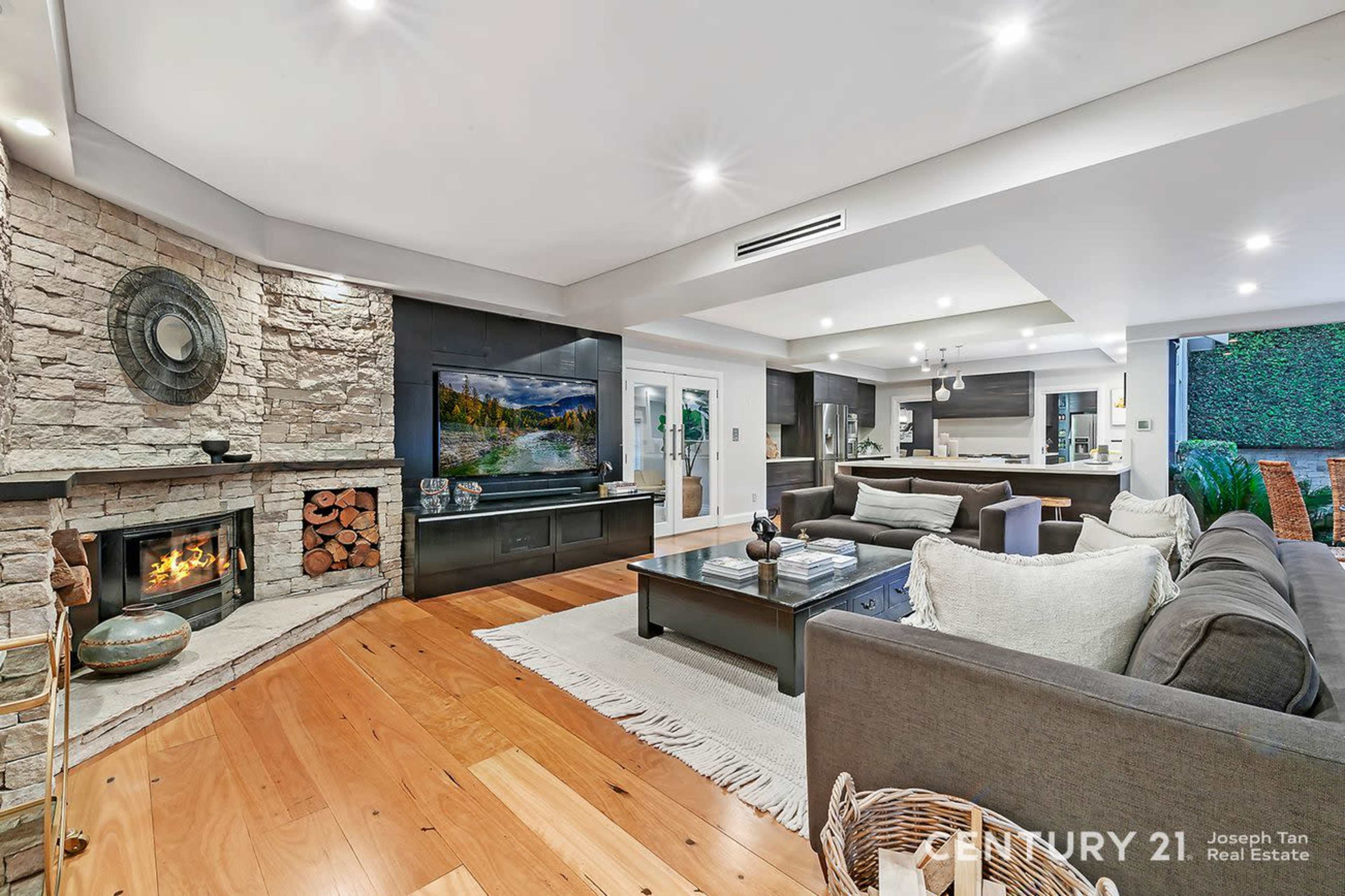 The image shows a modern living room featuring a stone fireplace, a large television mounted on the wall, and wooden flooring complemented by a dark sectional sofa and a coffee table.