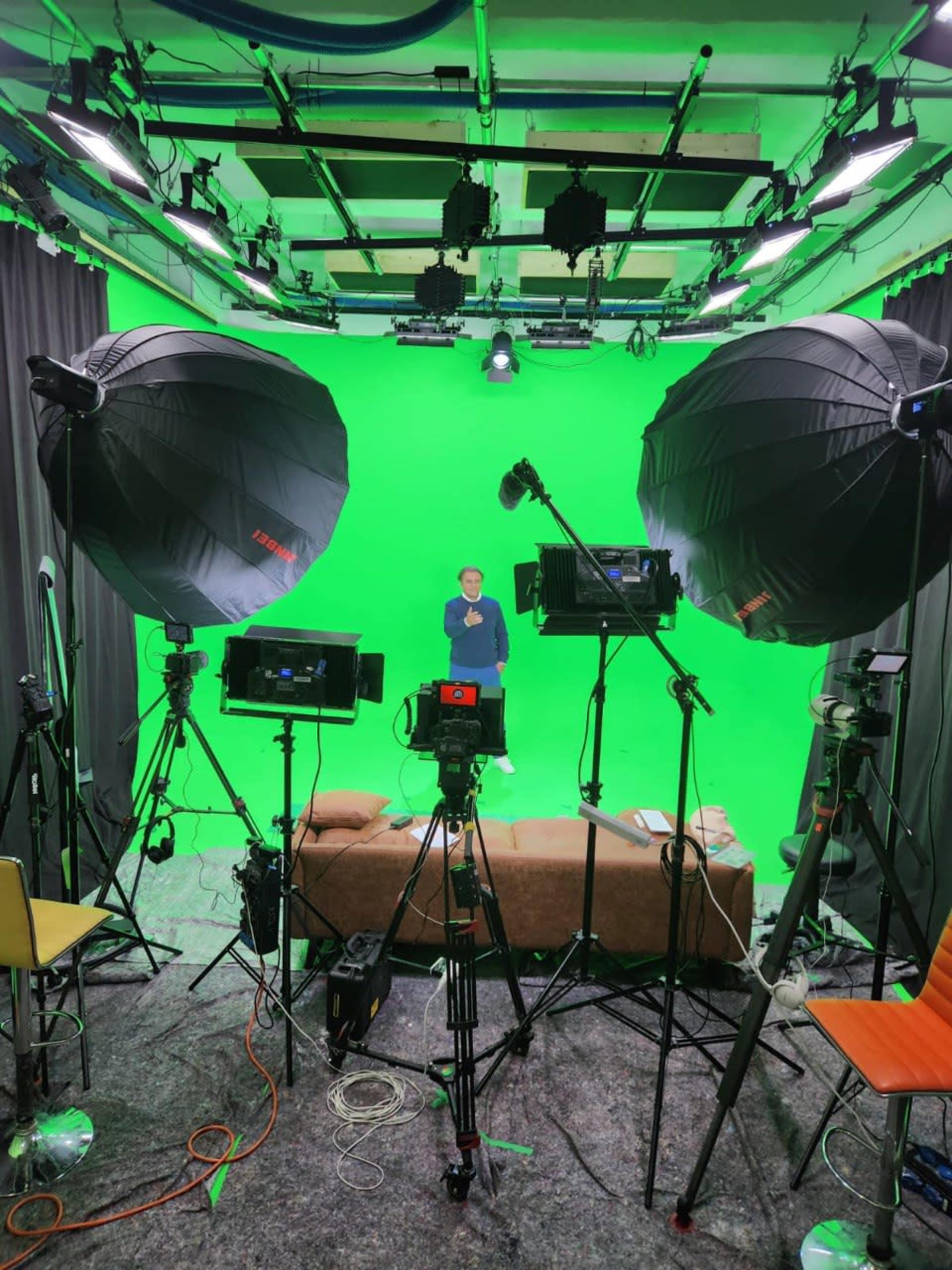A person stands in front of a green screen surrounded by studio lights and cameras.