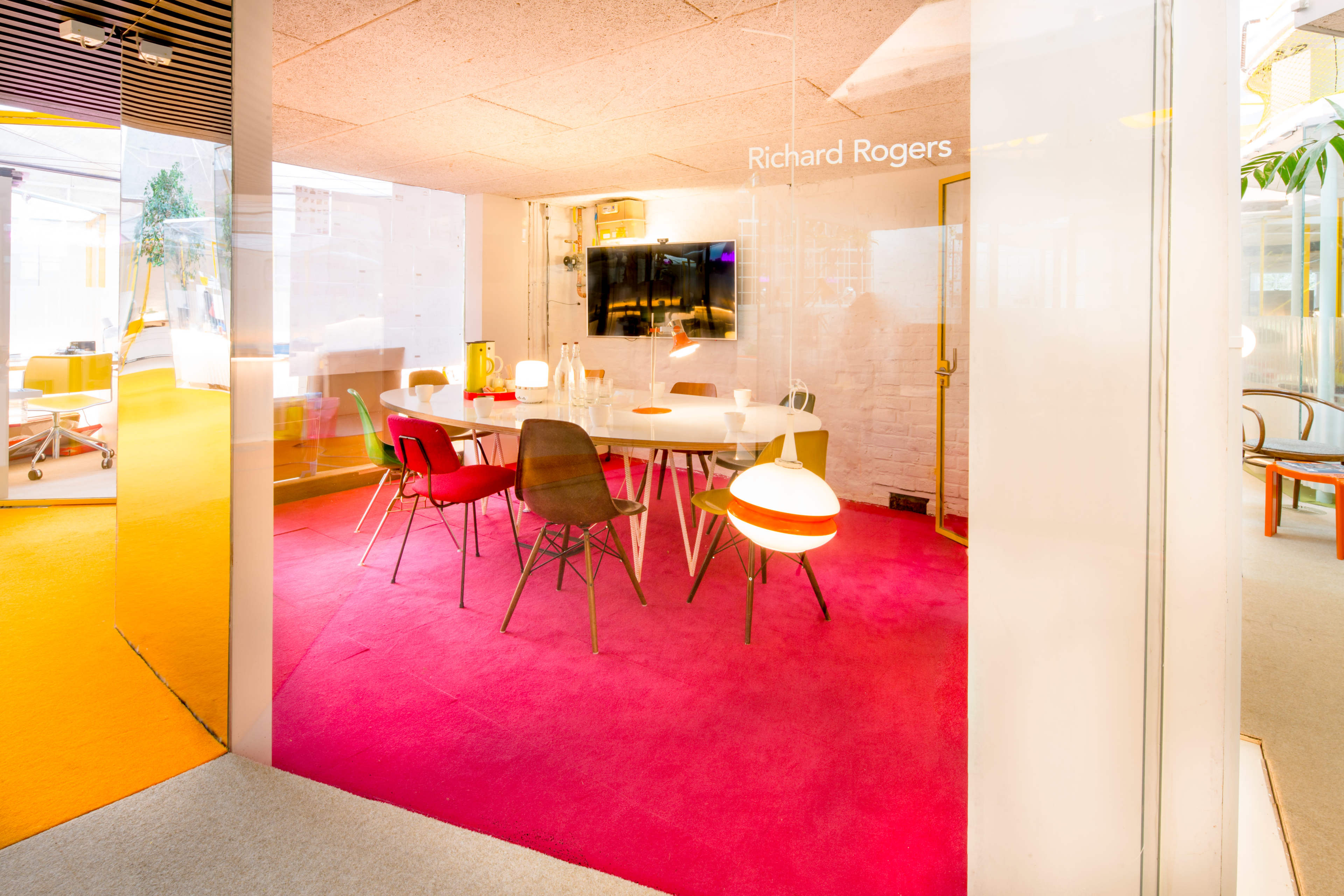 The image shows a modern office space featuring a glass-walled meeting room with a round table surrounded by chairs, set against a bright pink carpet.