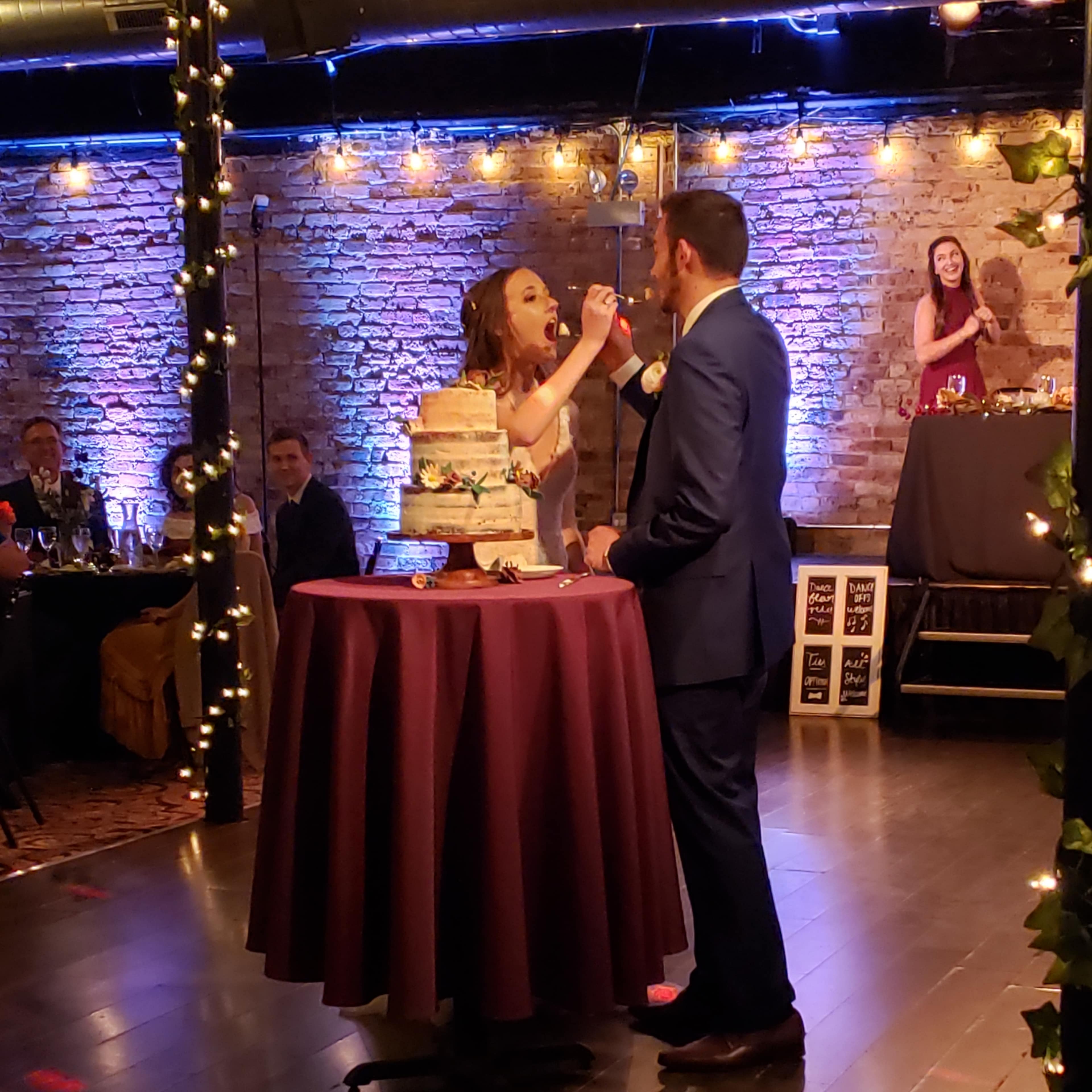 A woman feeds cake to a man while guests watch in a warmly lit venue with brick walls and festive decorations.