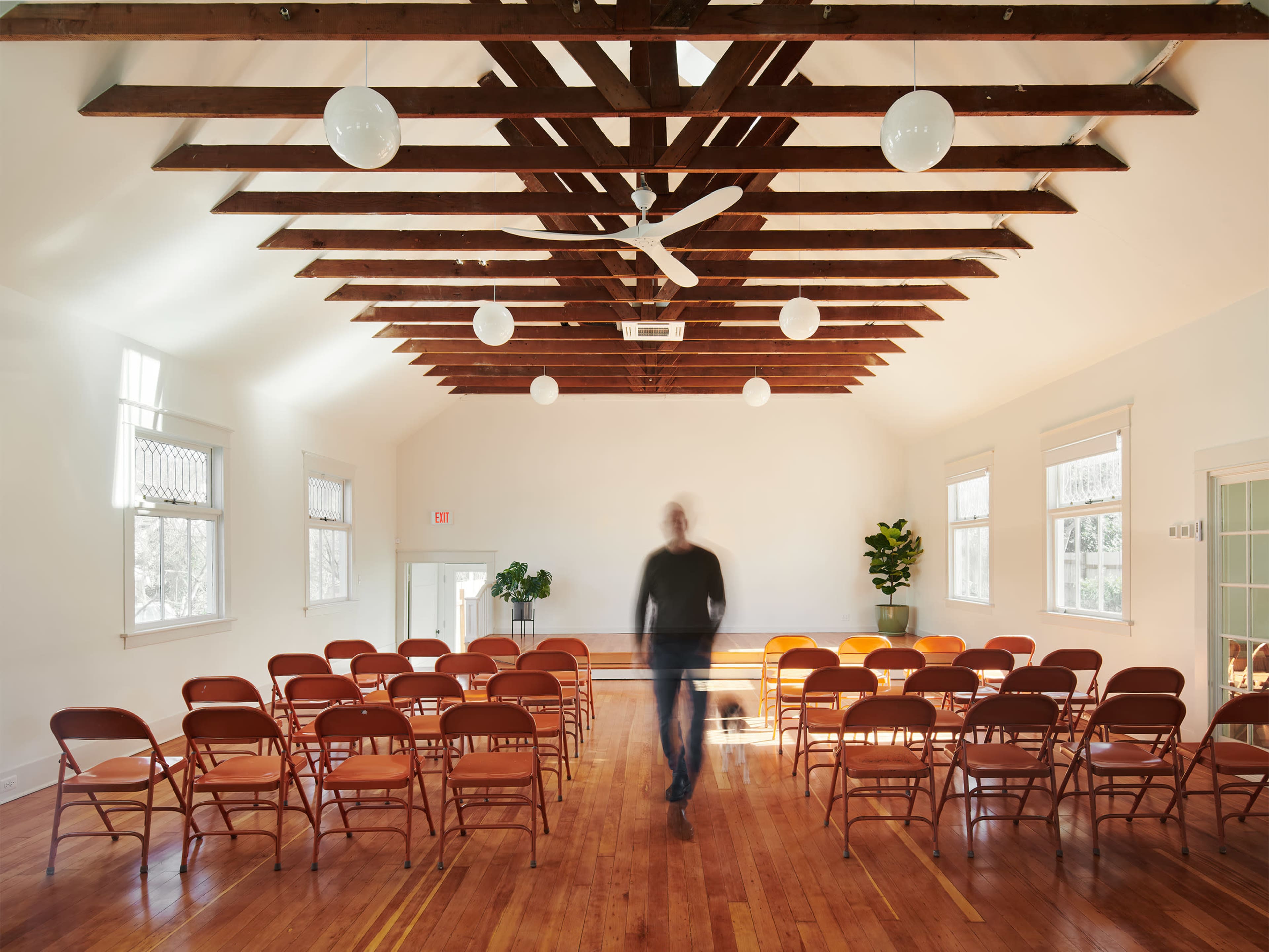 A man walks through a spacious, empty room with wooden beams, arranged chairs, and a potted plant.