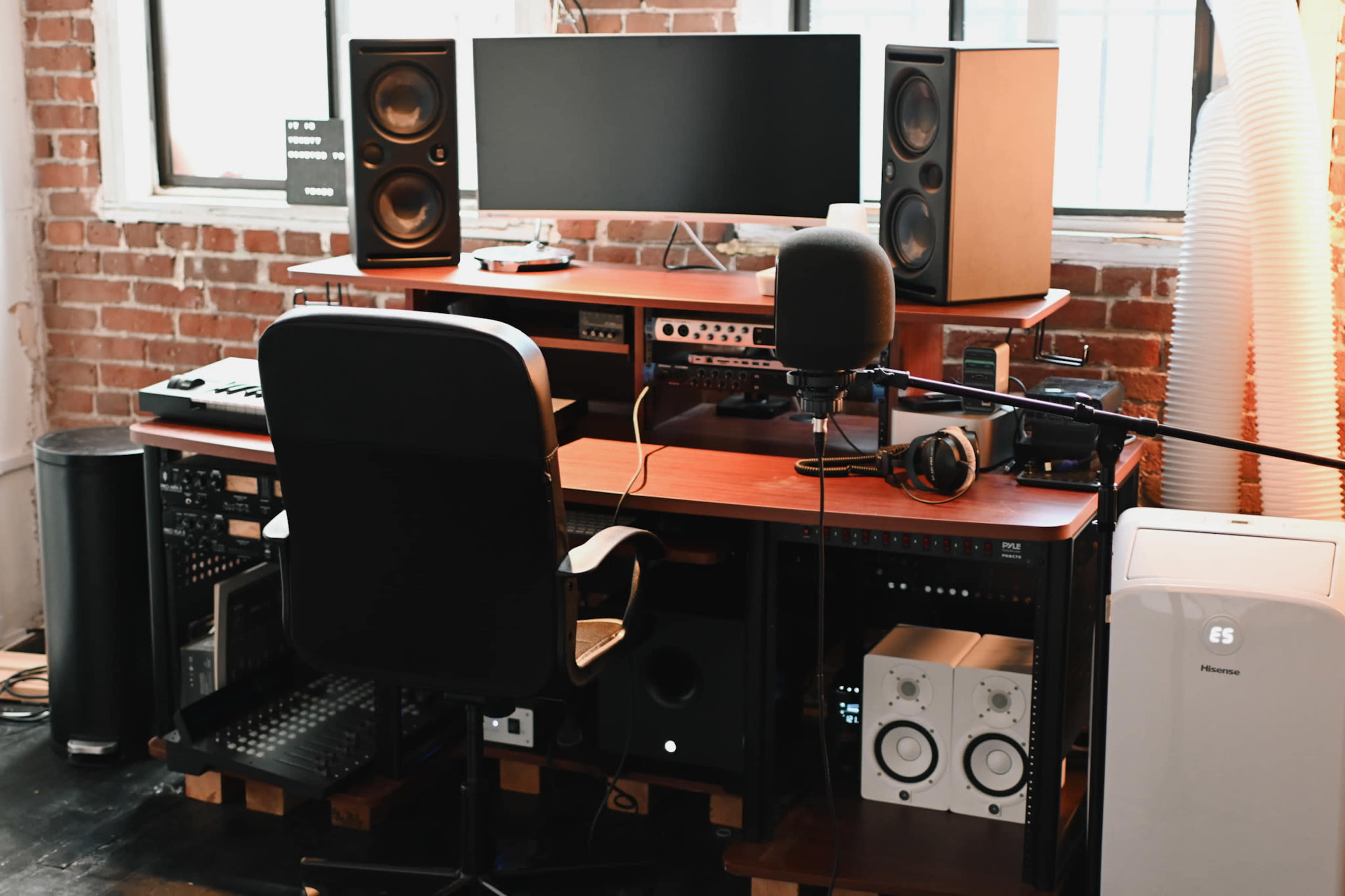 A music production workspace featuring a computer monitor, speakers, an audio interface, a microphone, headphones, and various audio equipment on a desk against a brick wall.