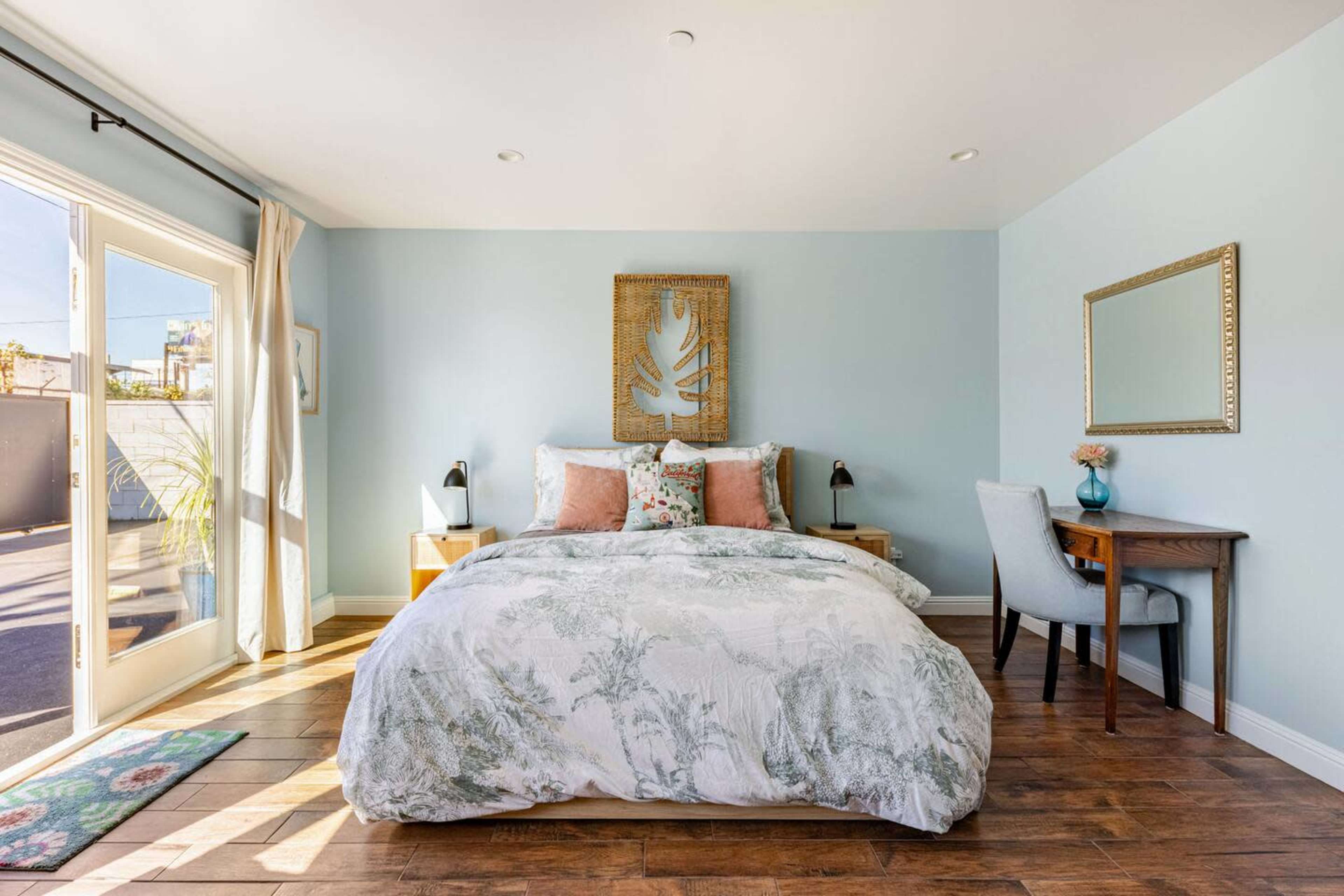 The bedroom features a queen-sized bed with a patterned comforter, two bedside tables, a desk, and large glass doors leading to an outdoor area.