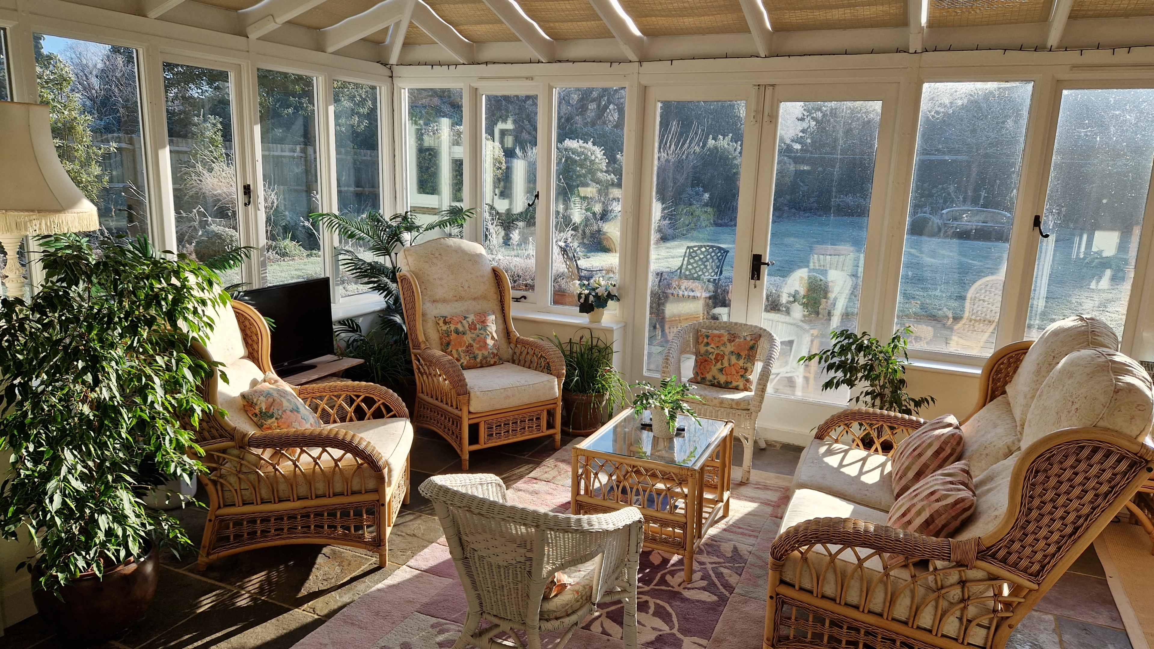 A sunlit conservatory features rattan furniture, including armchairs and a coffee table surrounded by indoor plants.