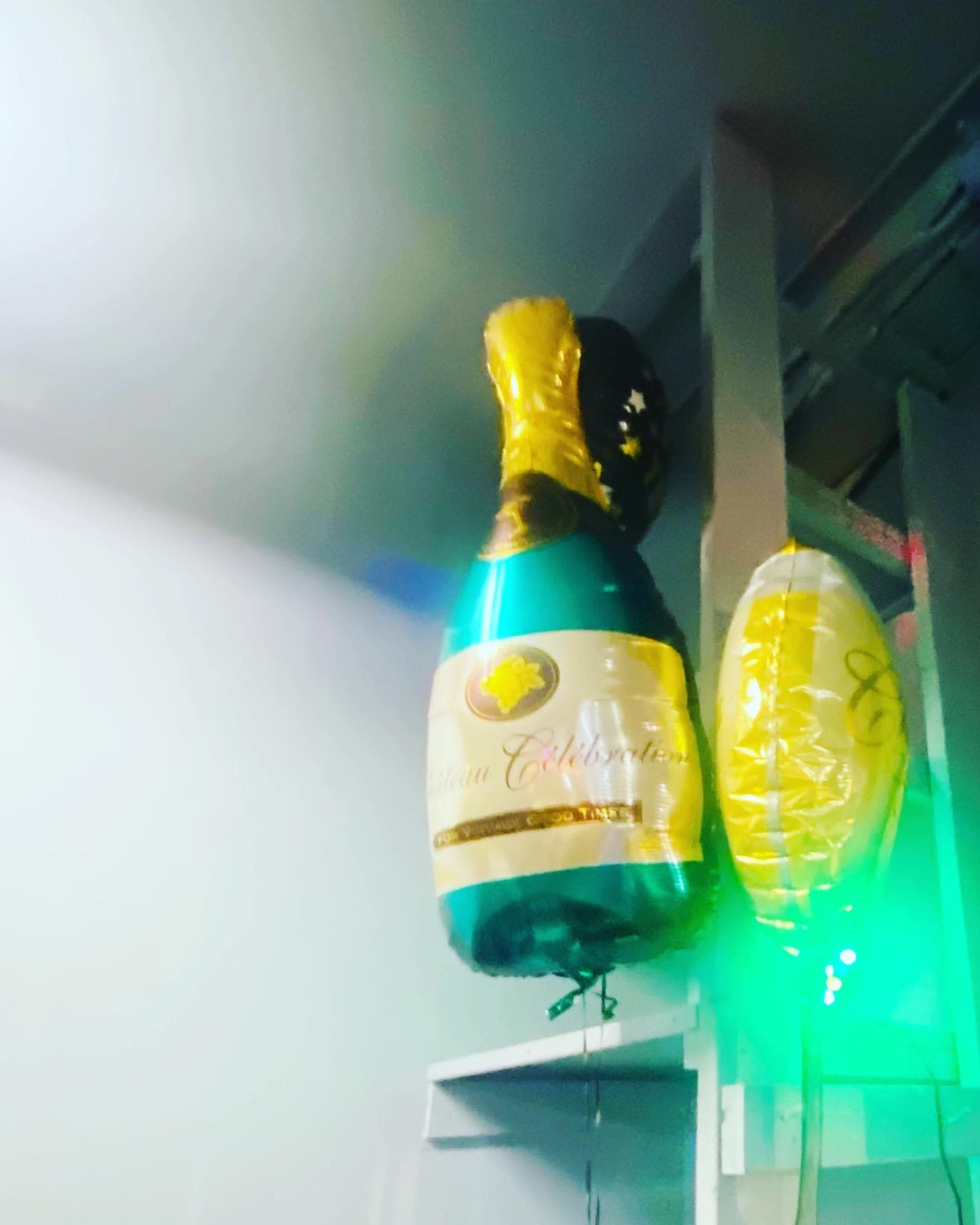 The image shows a helium-filled balloon designed to resemble a champagne bottle and another round yellow balloon on a wall.
