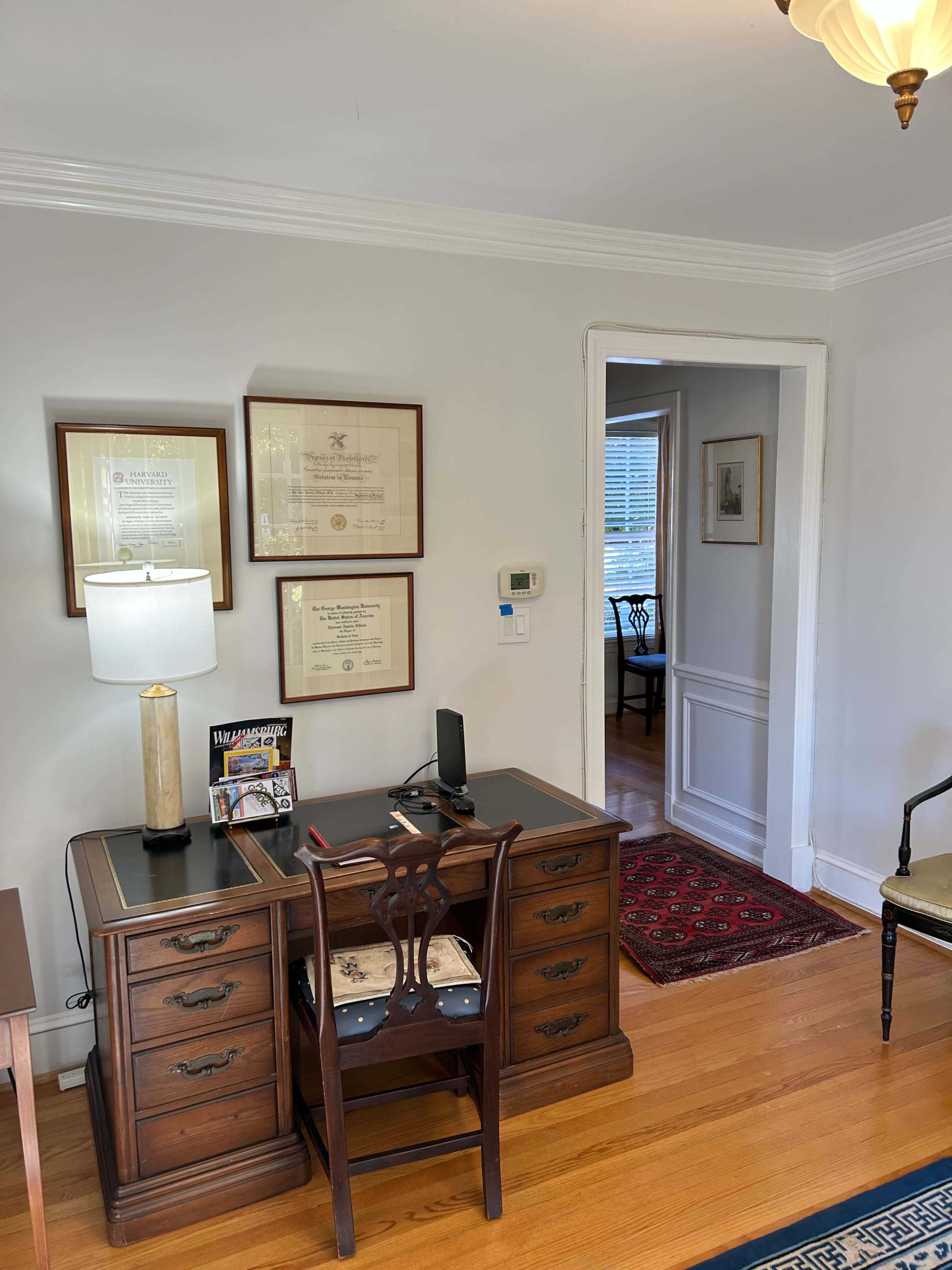 An office space features a wooden desk with a lamp, framed certificates on the wall, and a doorway leading to another room.