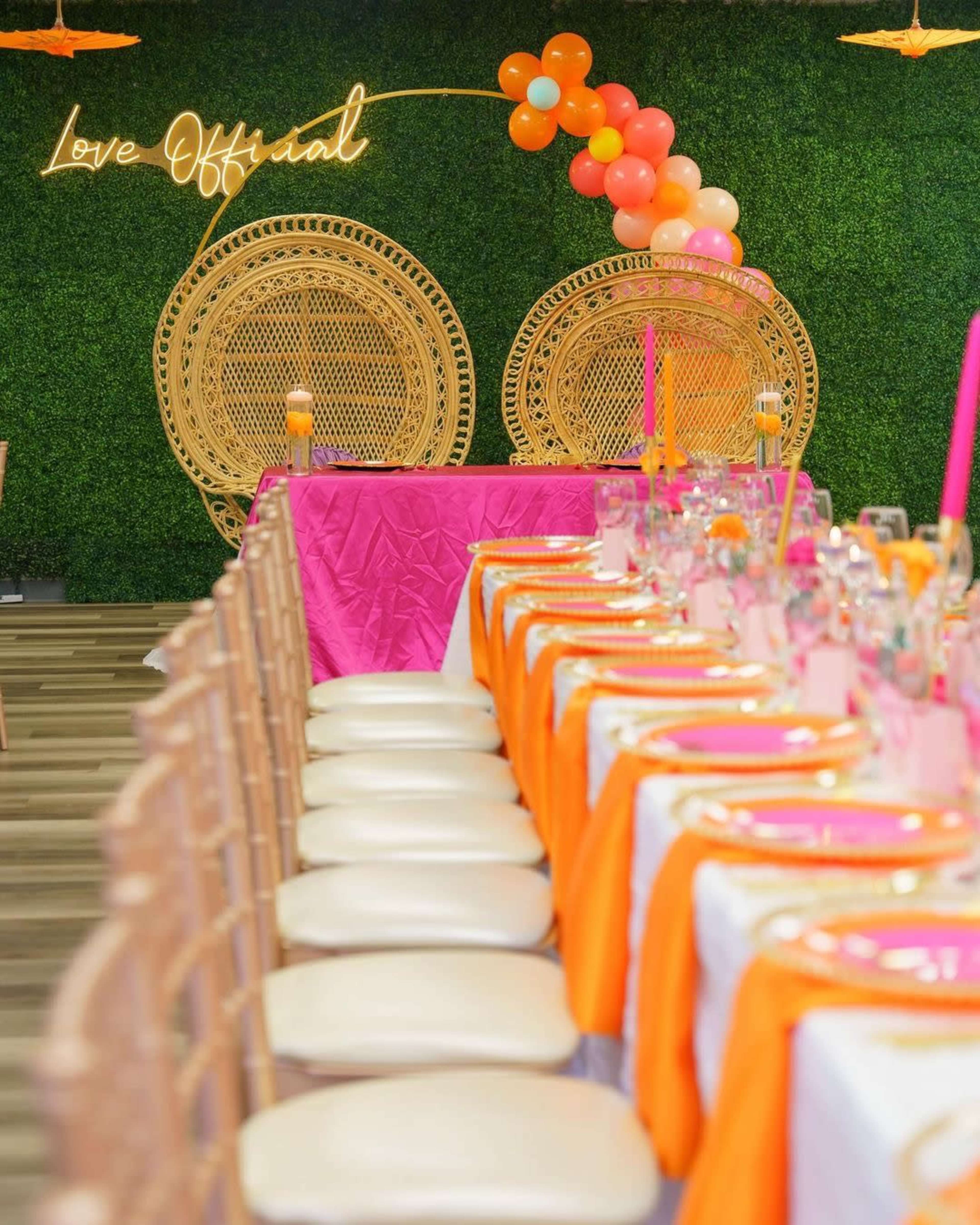 A decorated event space with a long table set with orange and pink tableware, flanked by two large wicker chairs and a neon sign that reads "Love Official."
