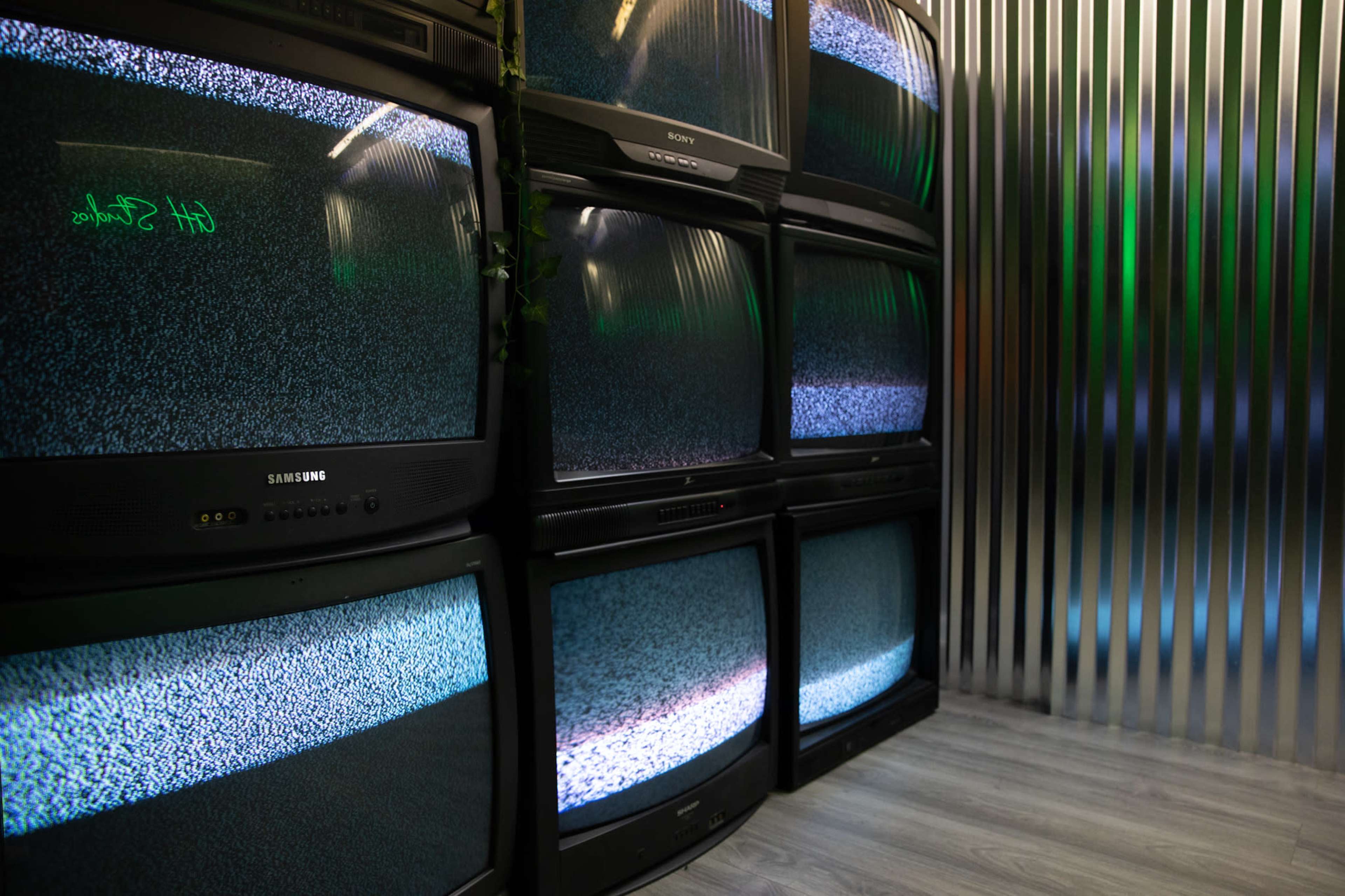 A stack of televisions displaying static is located against a metal-paneled wall.