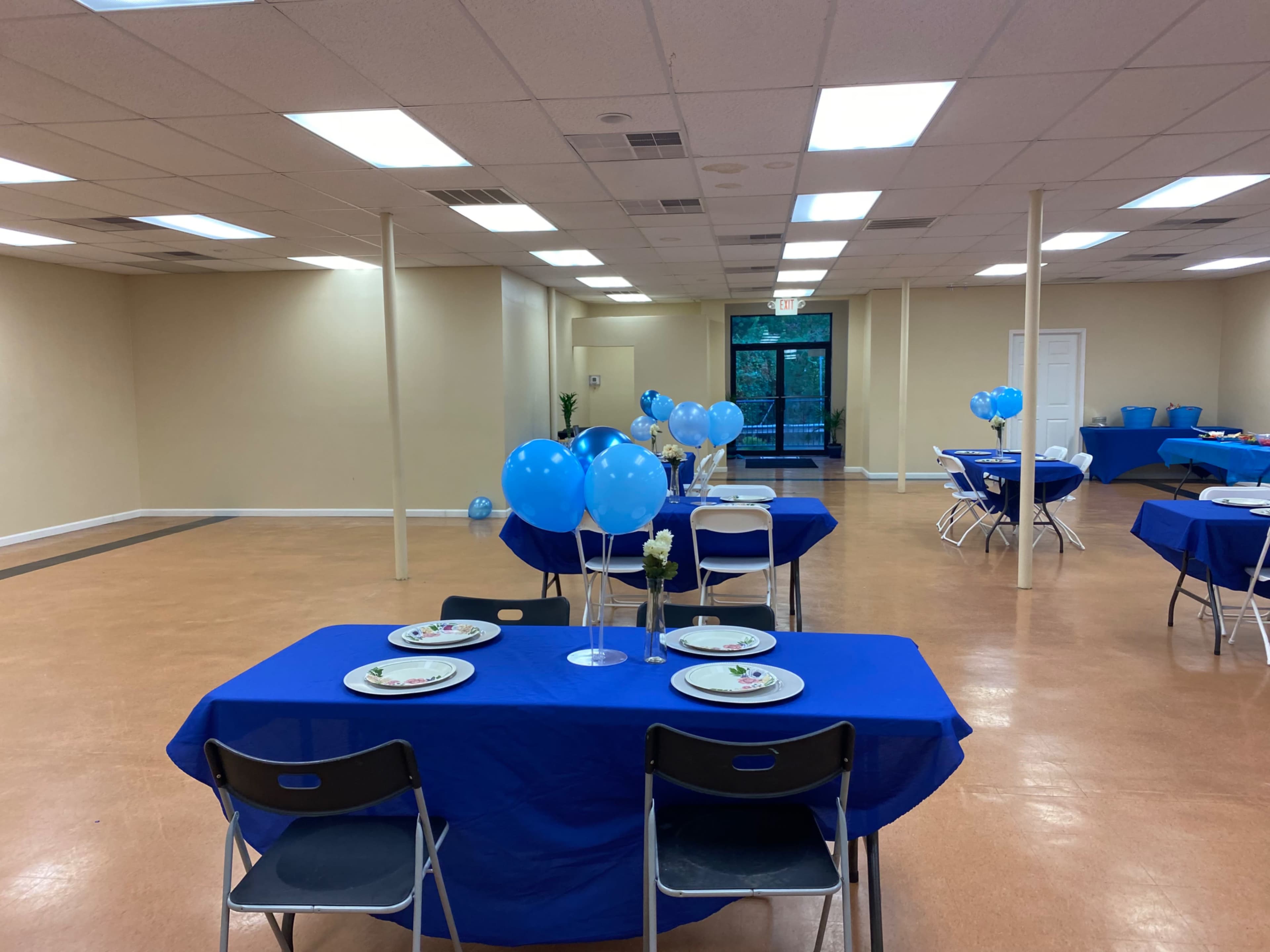 A large, empty room features several tables covered in blue cloths, set with plates and decorated with blue balloons.