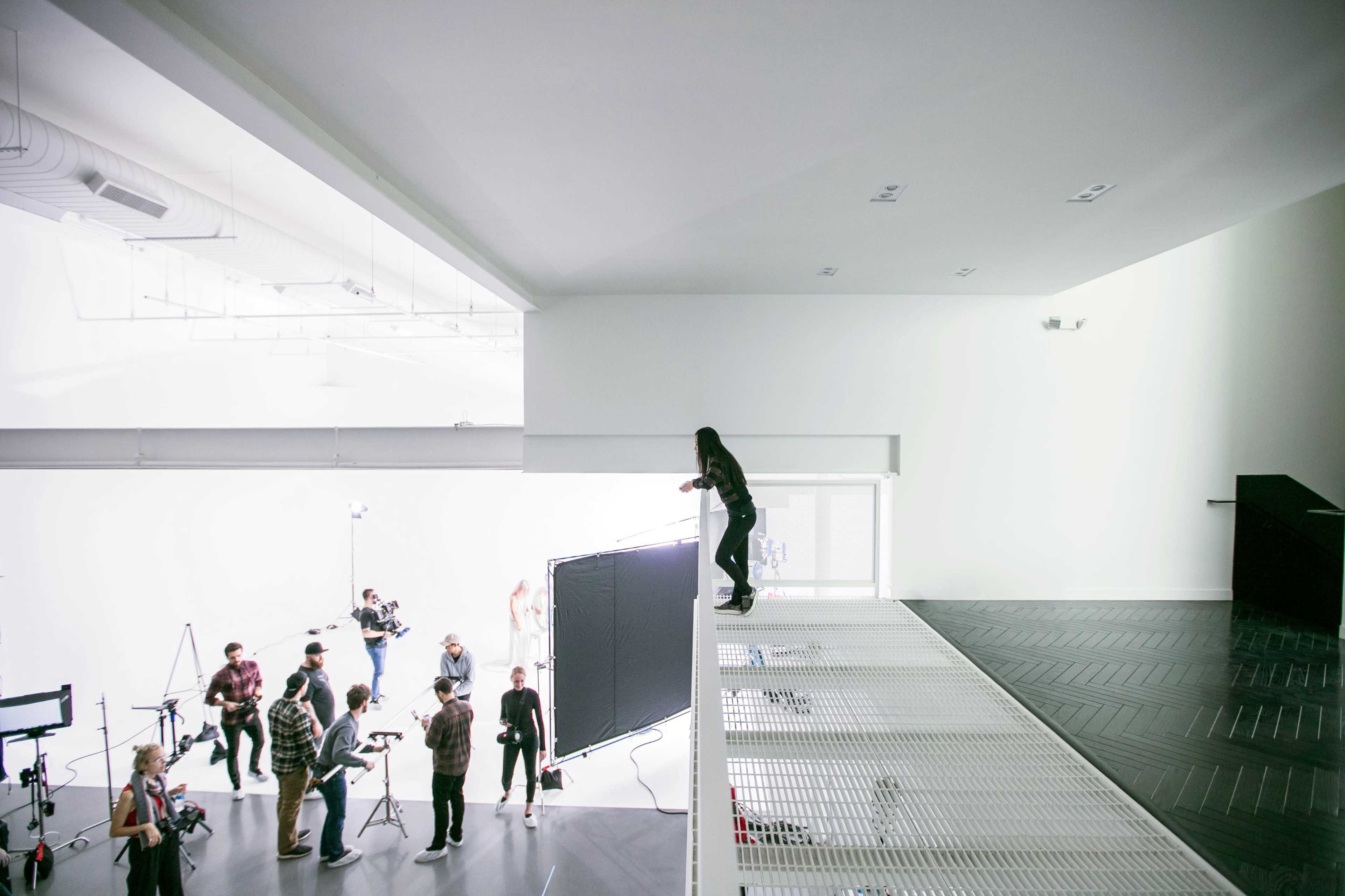 A person stands on a ledge overlooking a photoshoot in a bright studio with multiple crew members and equipment.