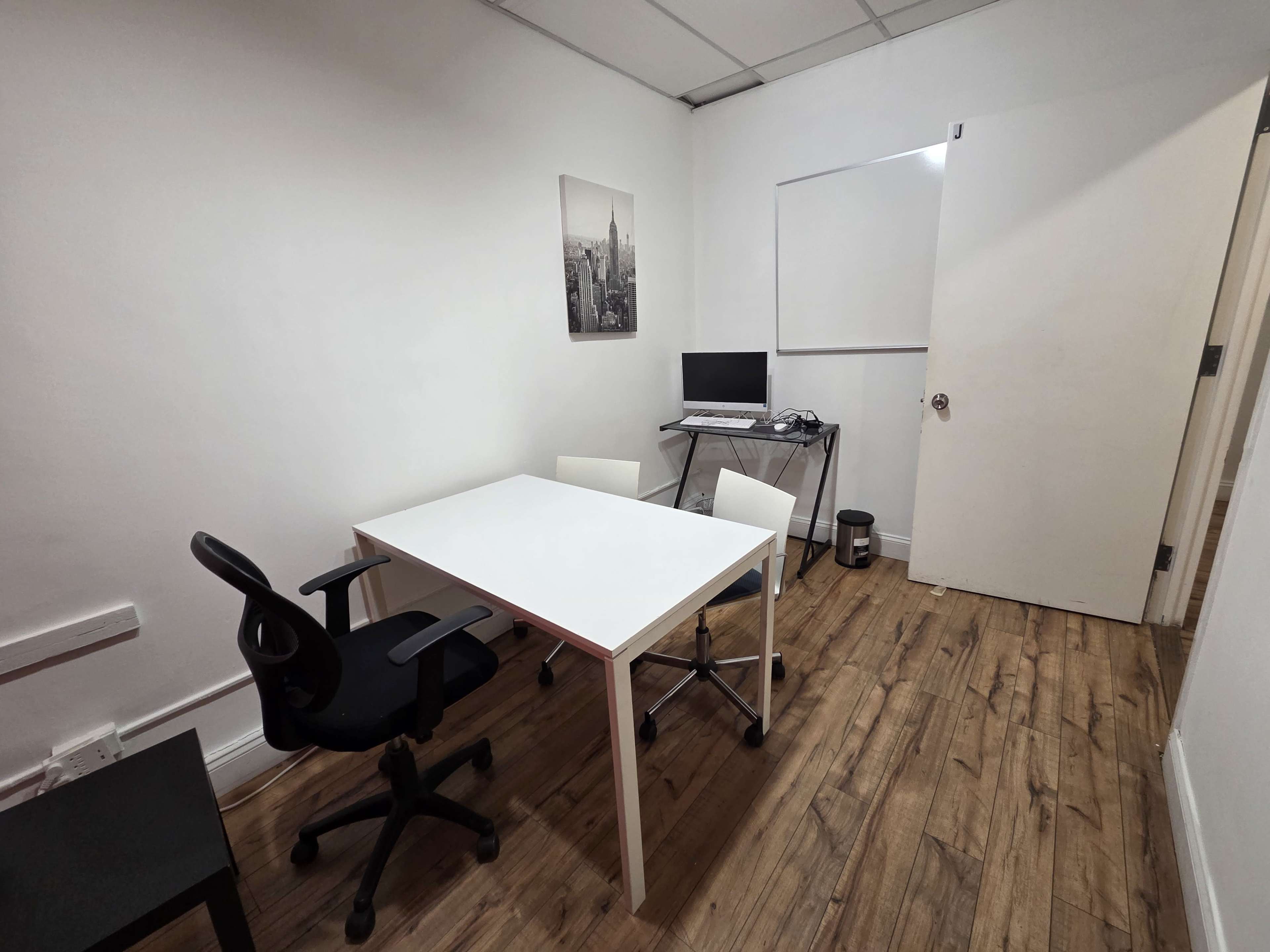 The image shows a small office with a white table and three chairs, a computer on a stand, and a whiteboard on the wall.