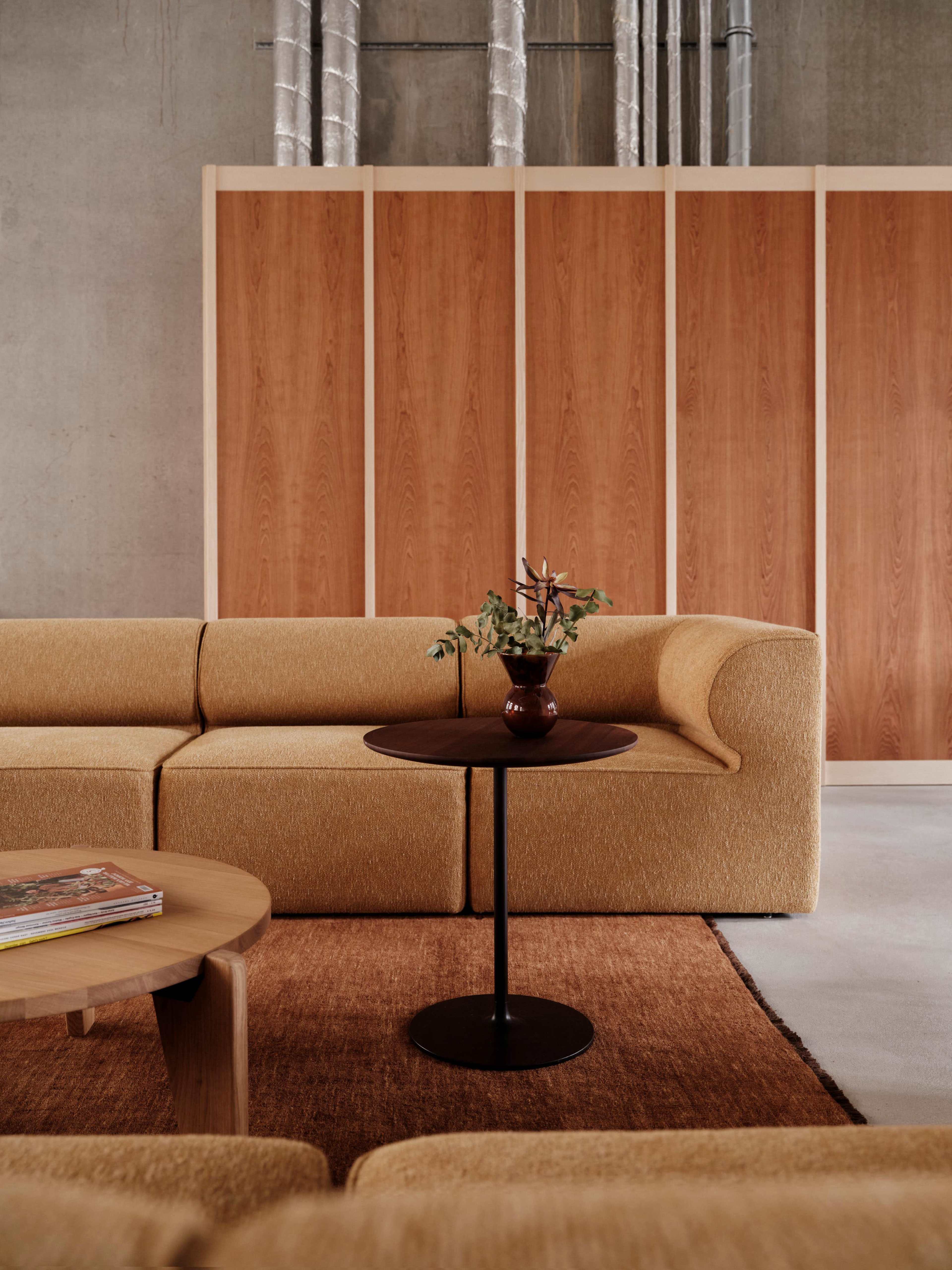 A modern living space features a beige sofa, a round black side table with a vase of greenery, and wooden partitions against a concrete wall.