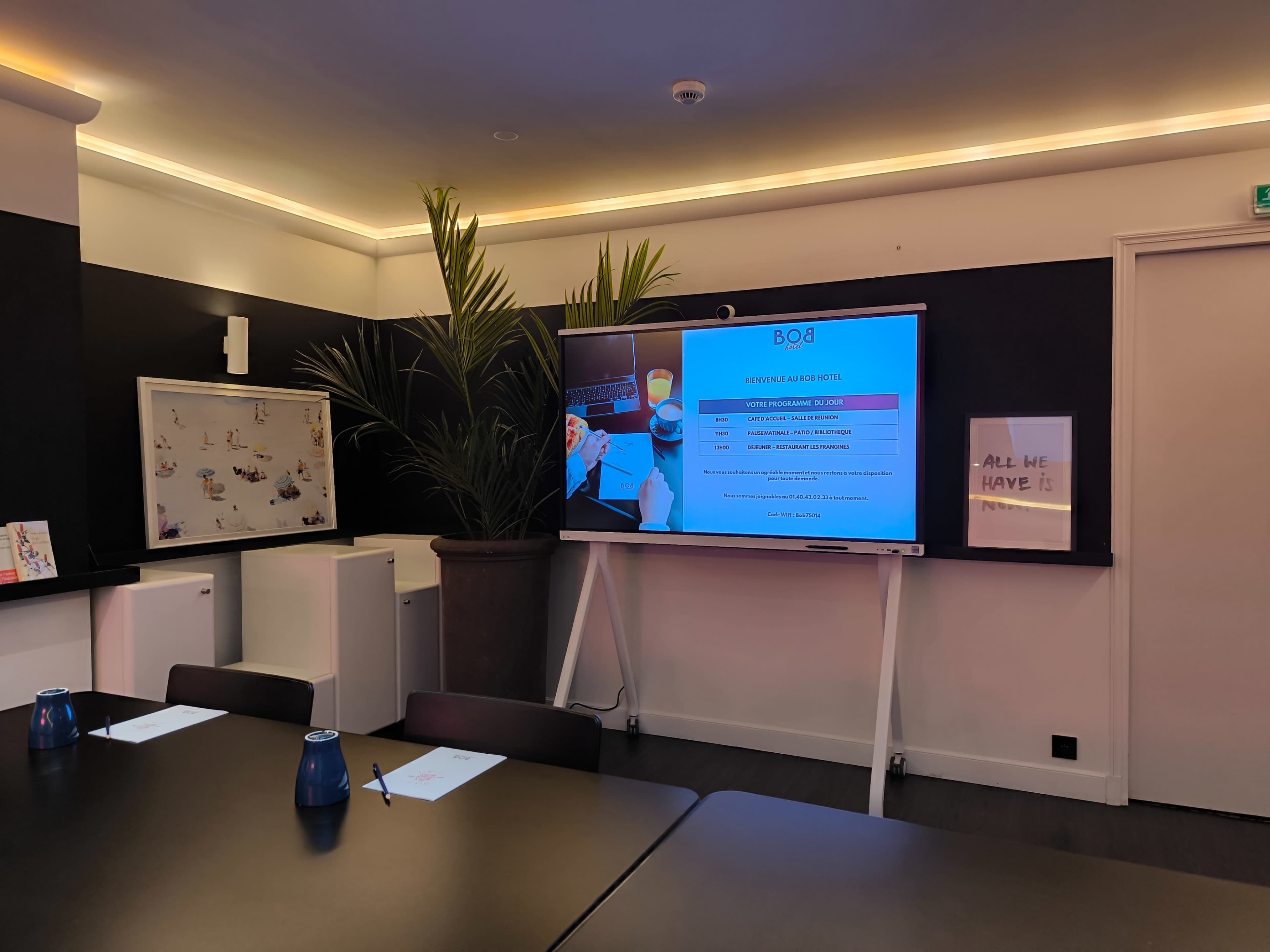 A meeting room with a large screen displaying information, a potted plant in the corner, and a conference table set with blue water glasses and notepads.