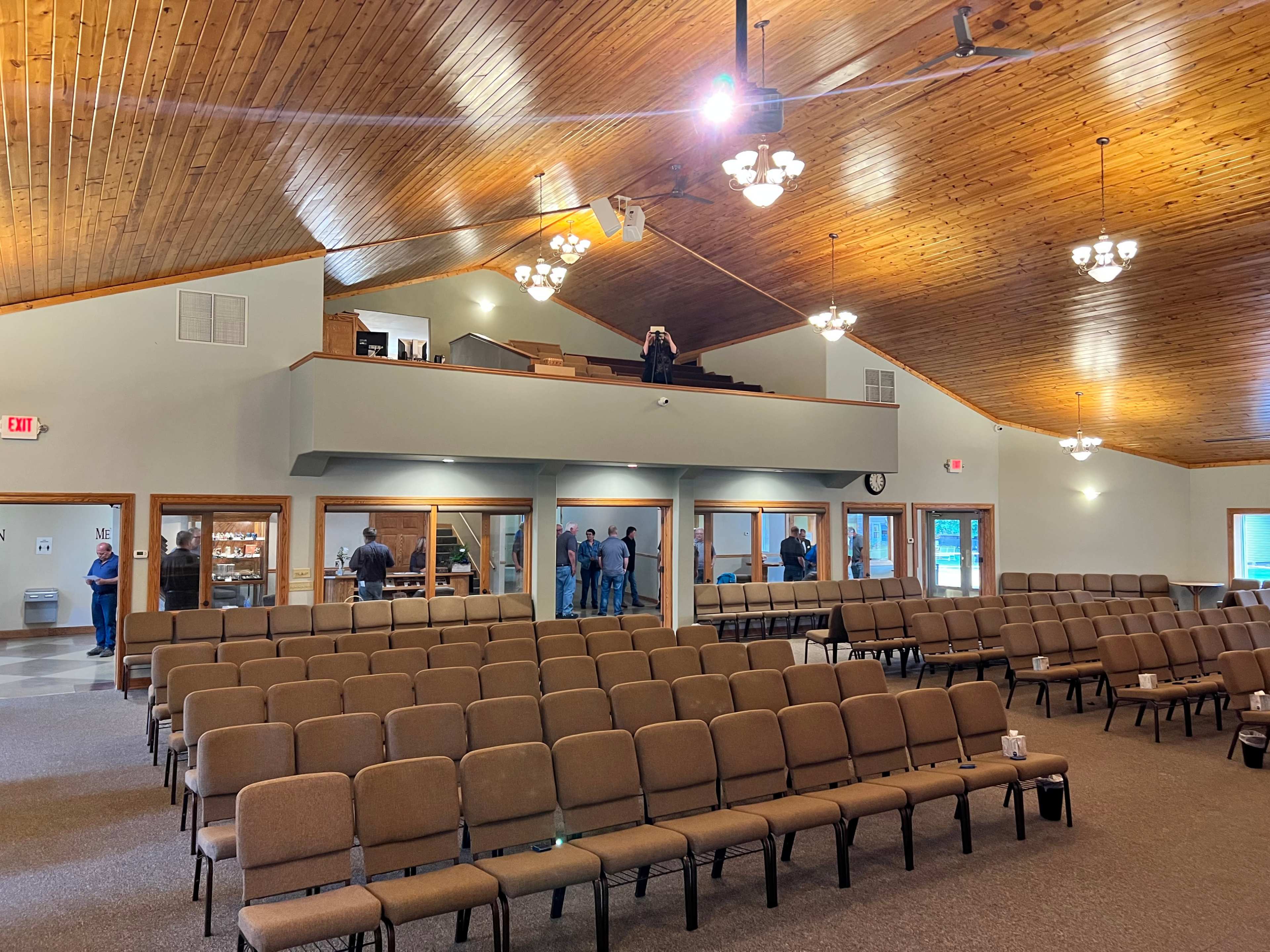 An interior view of a spacious auditorium with rows of brown chairs and a stage area, where a few people are gathered at the back.