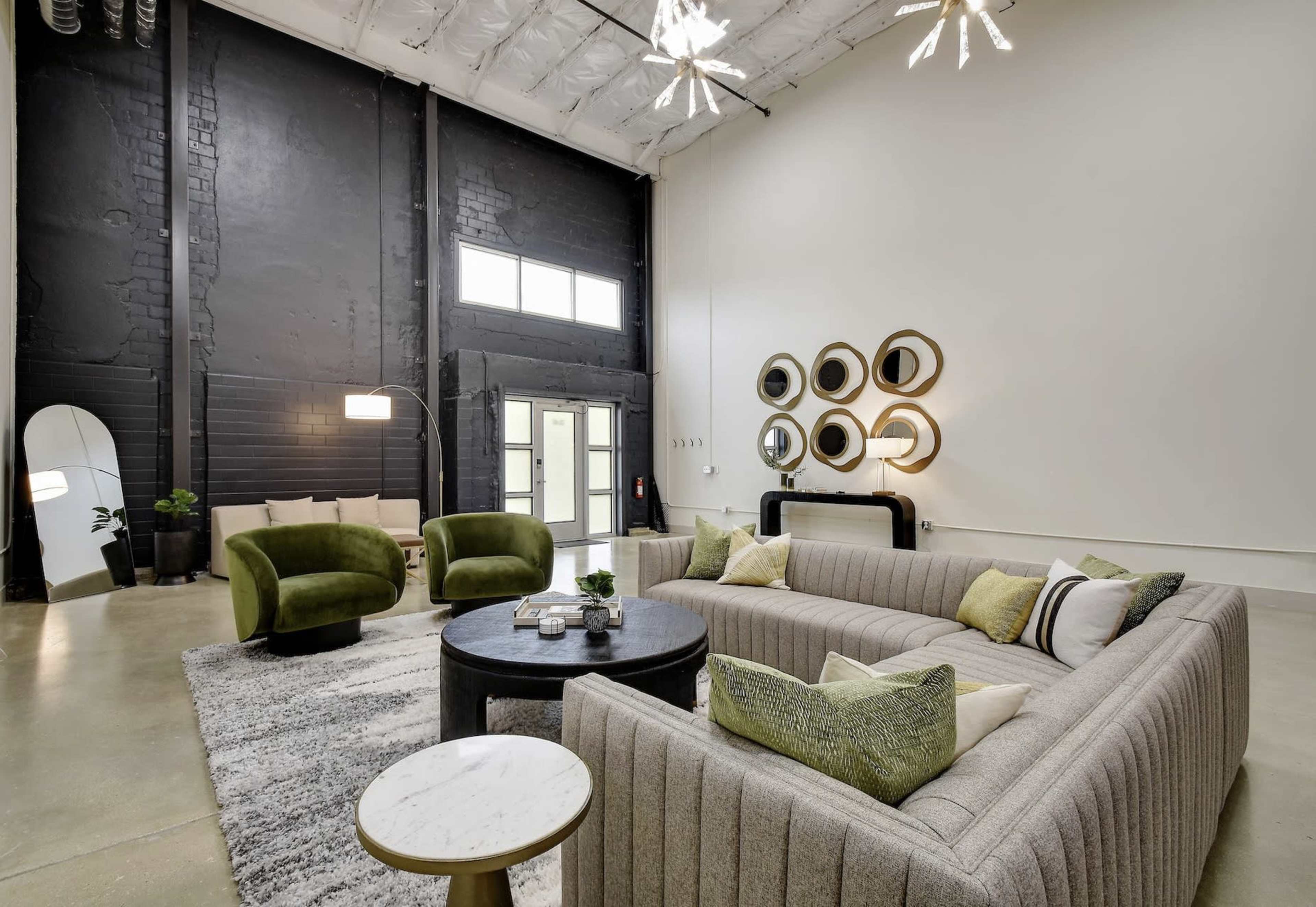 The image shows a modern, spacious living area with a large sectional sofa, two green armchairs, a round coffee table, and decorative mirrors on the wall.
