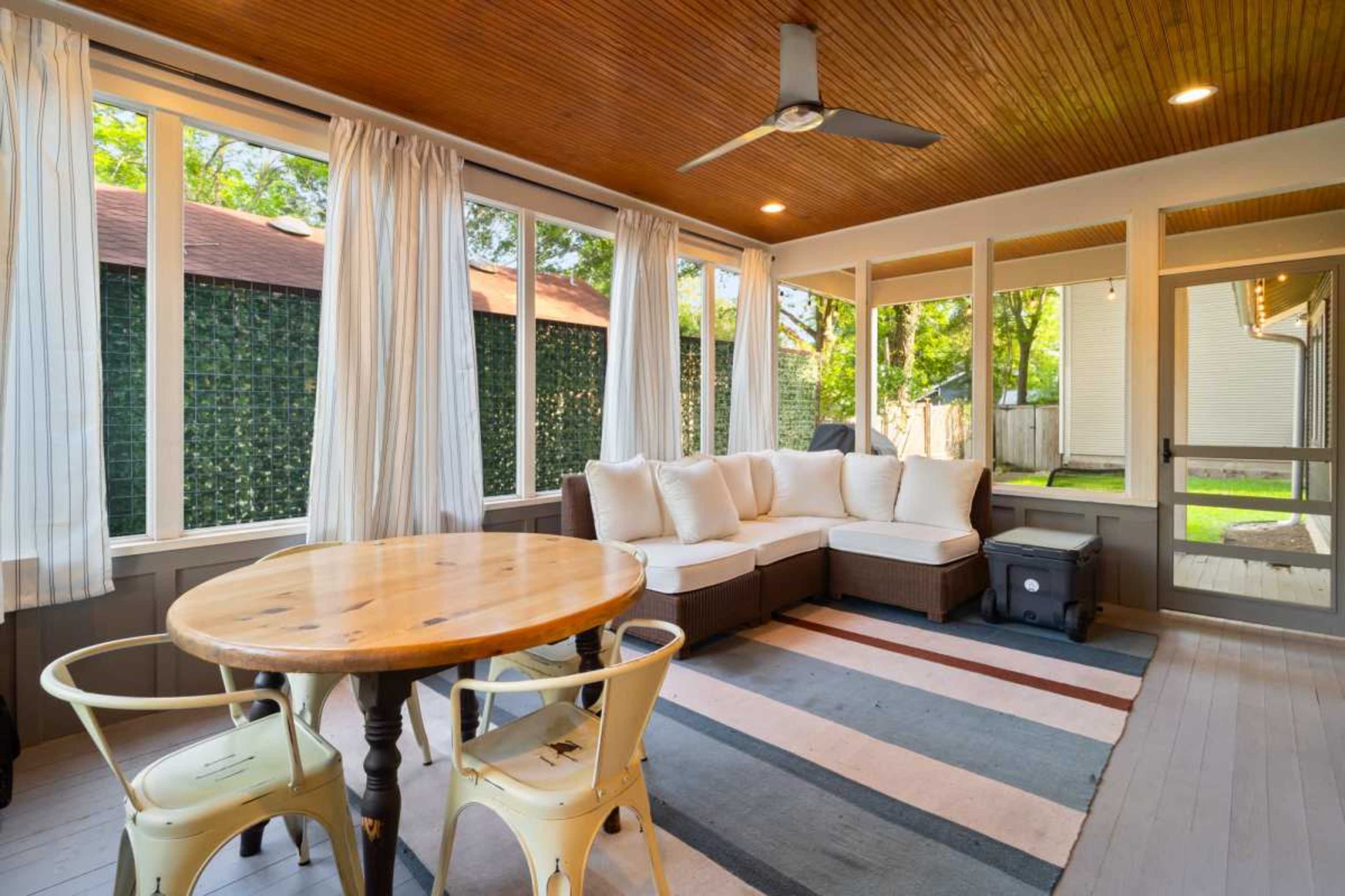 The image shows a bright, screened-in porch featuring a circular wooden table, two metal chairs, and a sectional sofa with light cushions, surrounded by large windows.