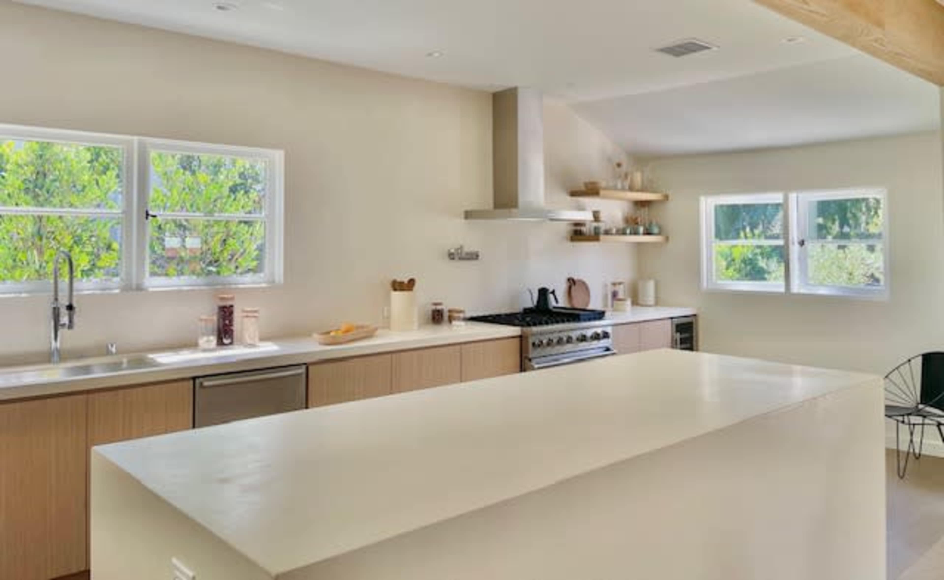 The image shows a modern kitchen with a large island, light wood cabinetry, and a gas stove against a neutral-colored wall, illuminated by natural light from the windows.