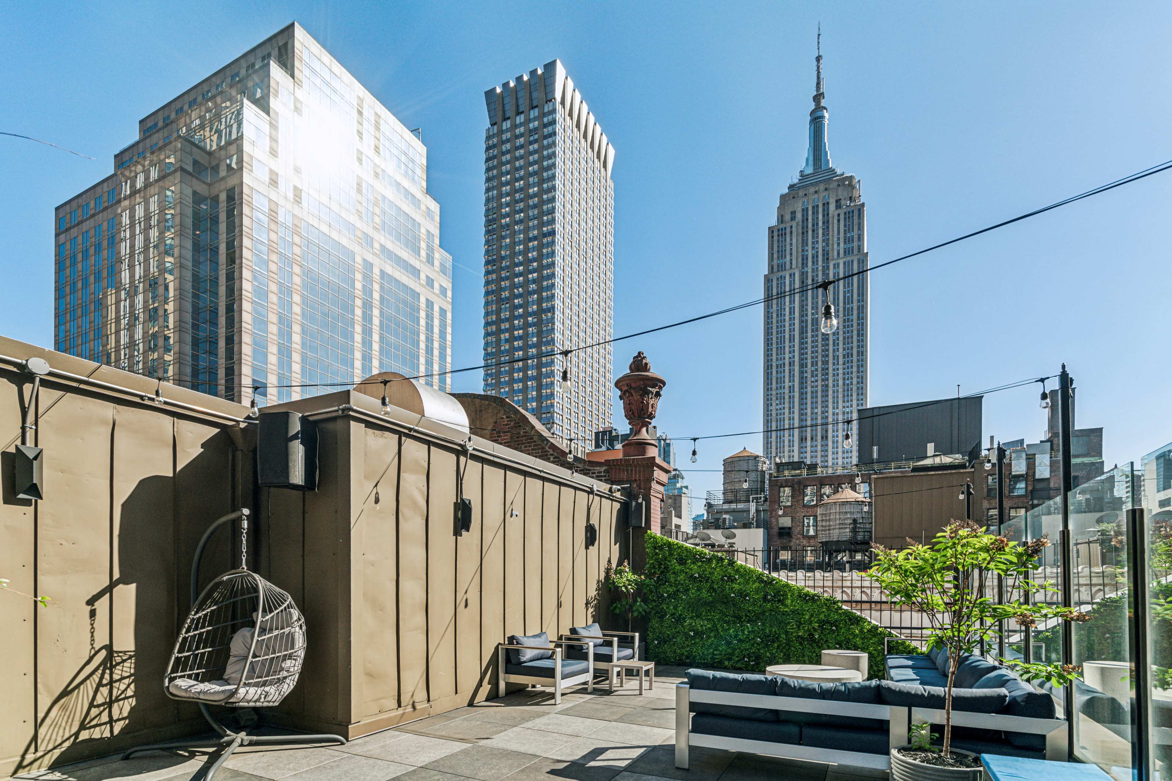 A rooftop terrace with modern seating and a view of the Empire State Building and surrounding skyscrapers under a clear blue sky.