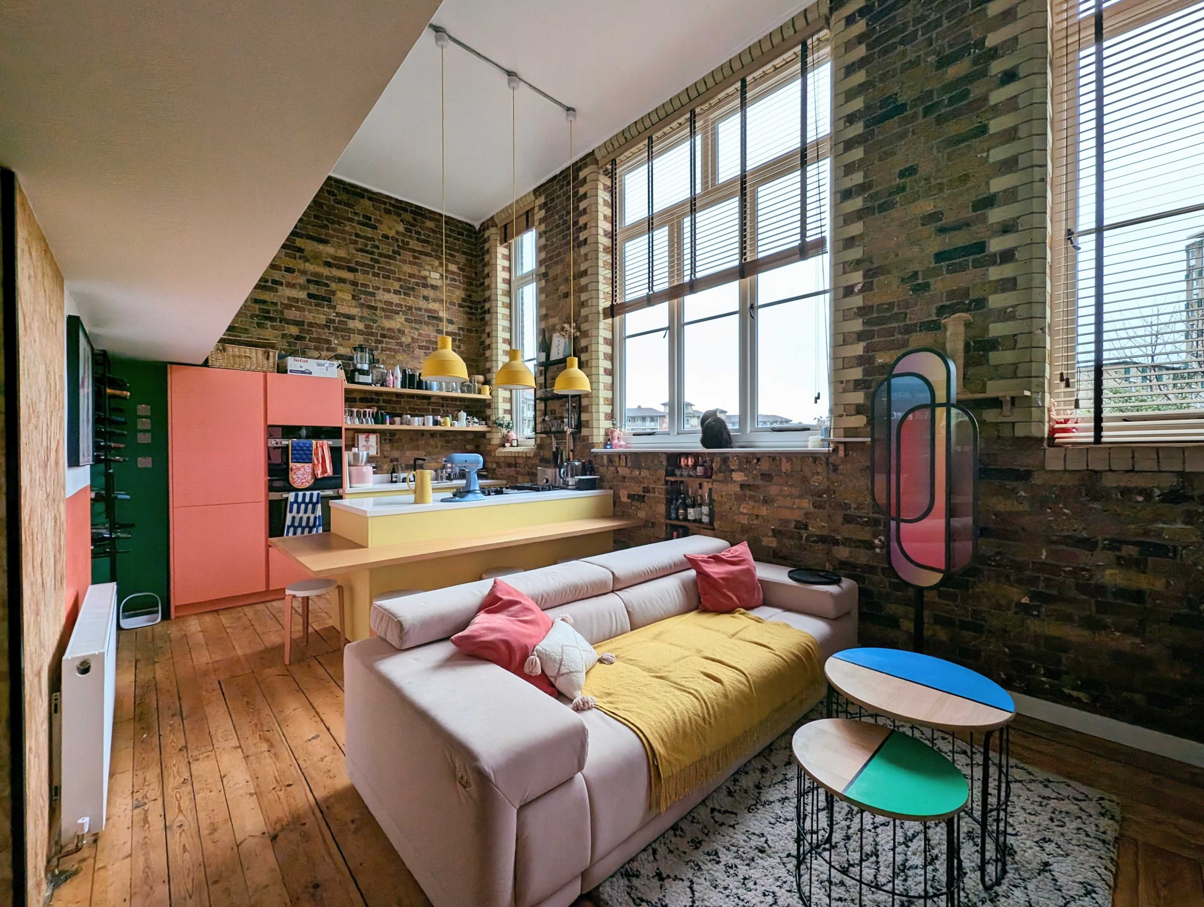 A modern open-plan living space with exposed brick walls, a sofa with colorful cushions, a kitchen with pink cabinets, and large windows letting in natural light.