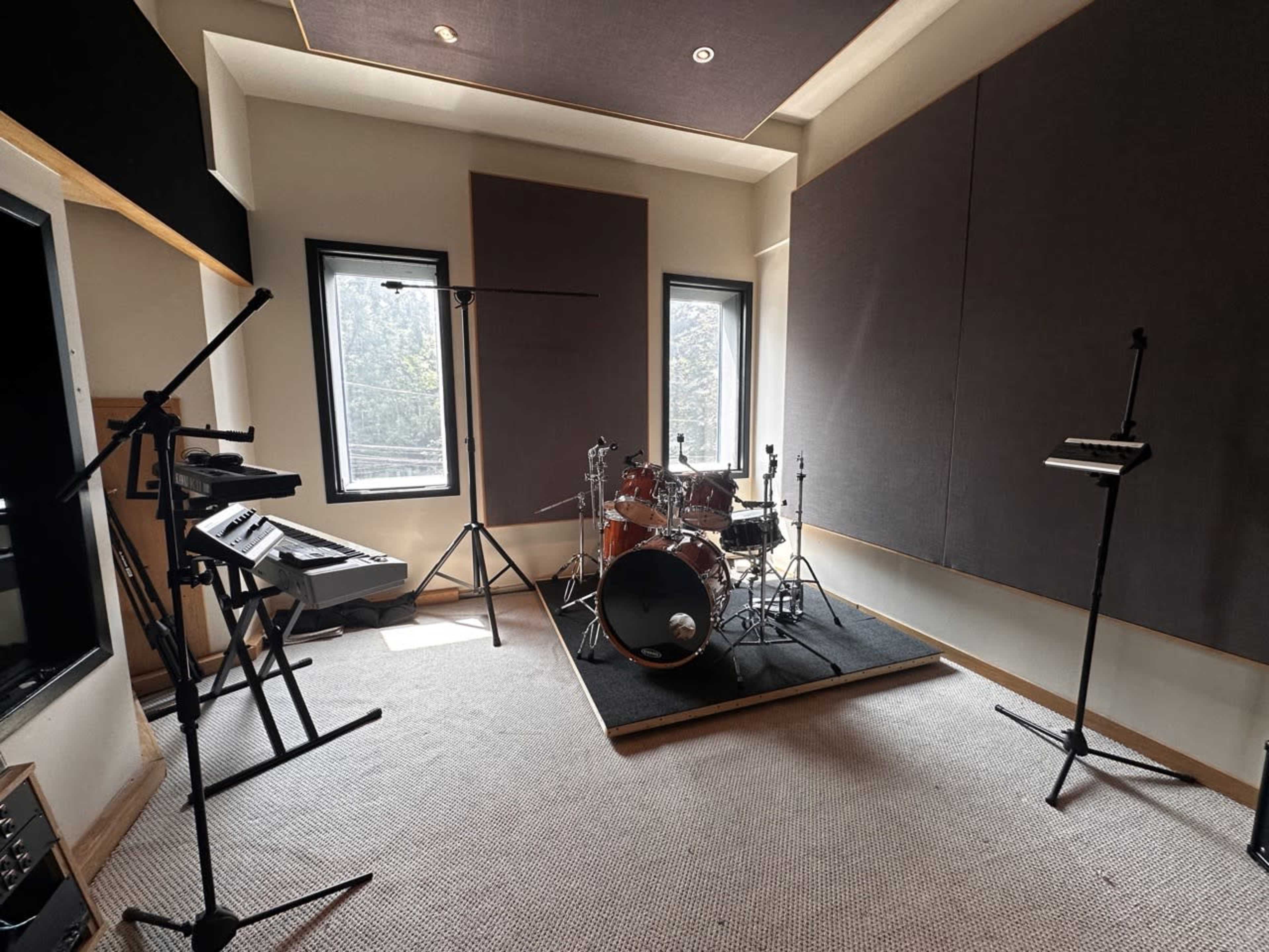 The image shows a soundproof music studio featuring a drum kit, keyboard, and microphone stands positioned in a well-lit space with large windows.