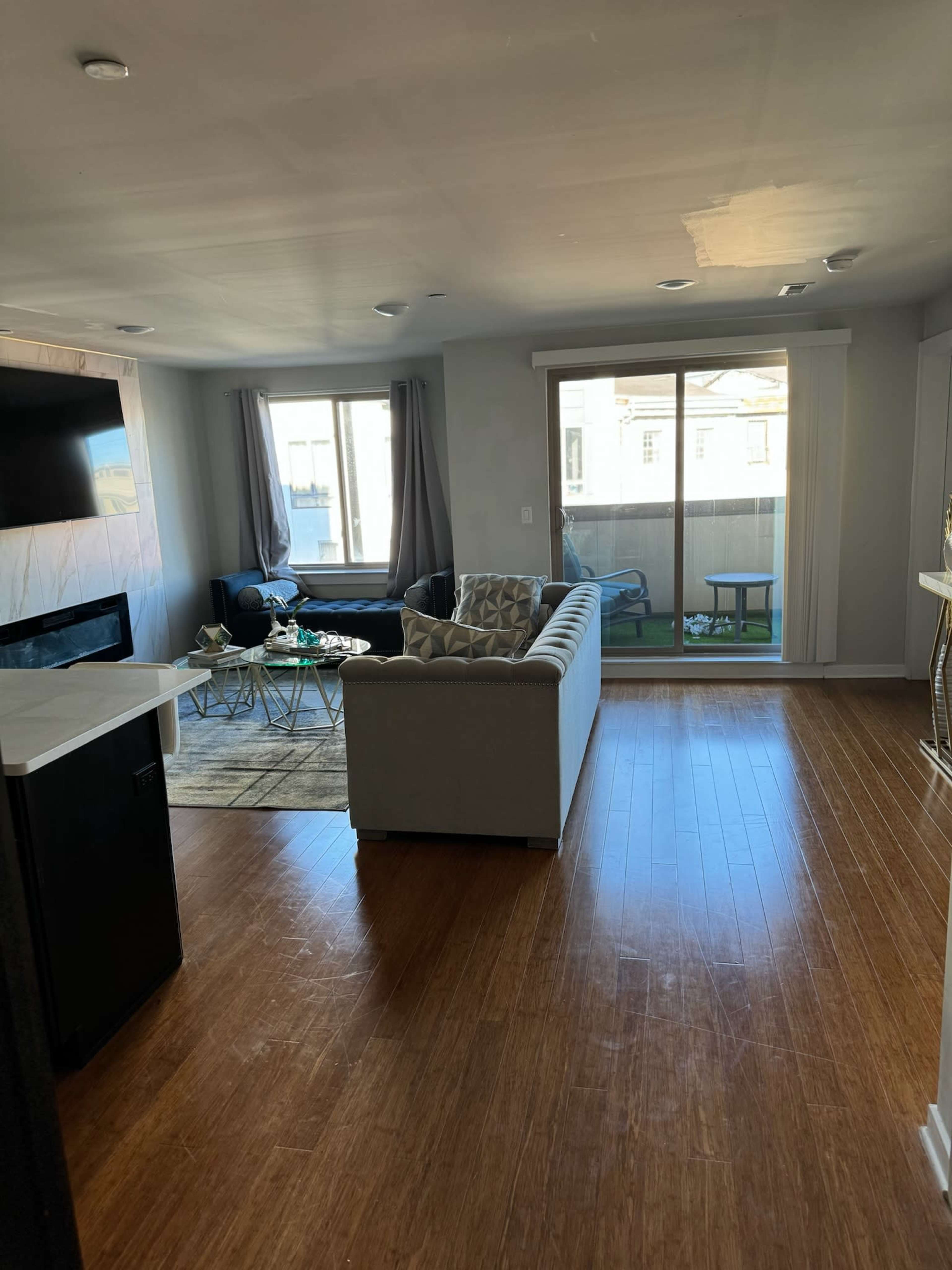 The image shows a modern, open-concept living room with hardwood floors, a large window with natural light, and a couch facing a coffee table.