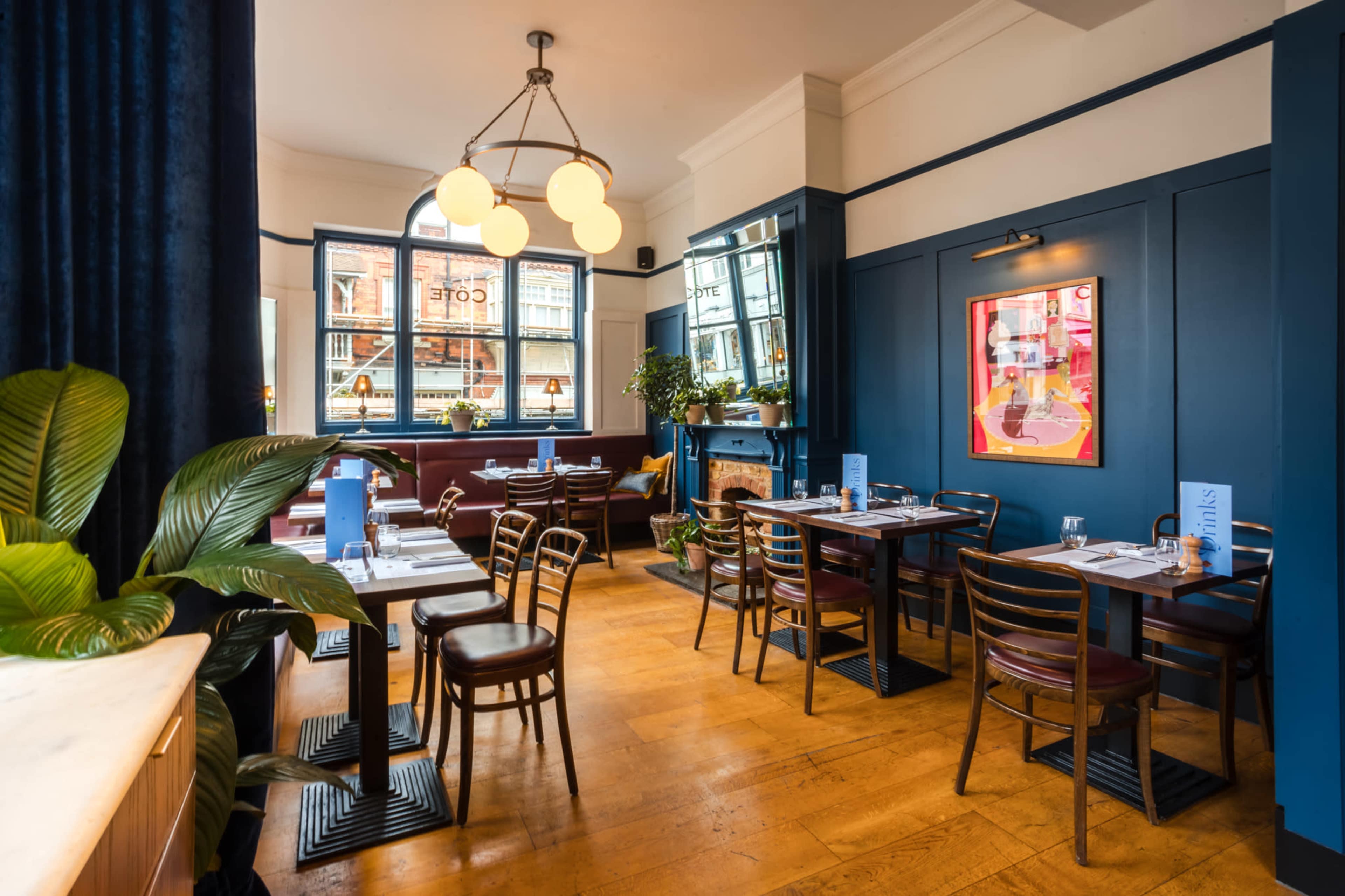The image shows a restaurant interior featuring wooden tables and chairs, blue walls, and large windows with decorative plants.