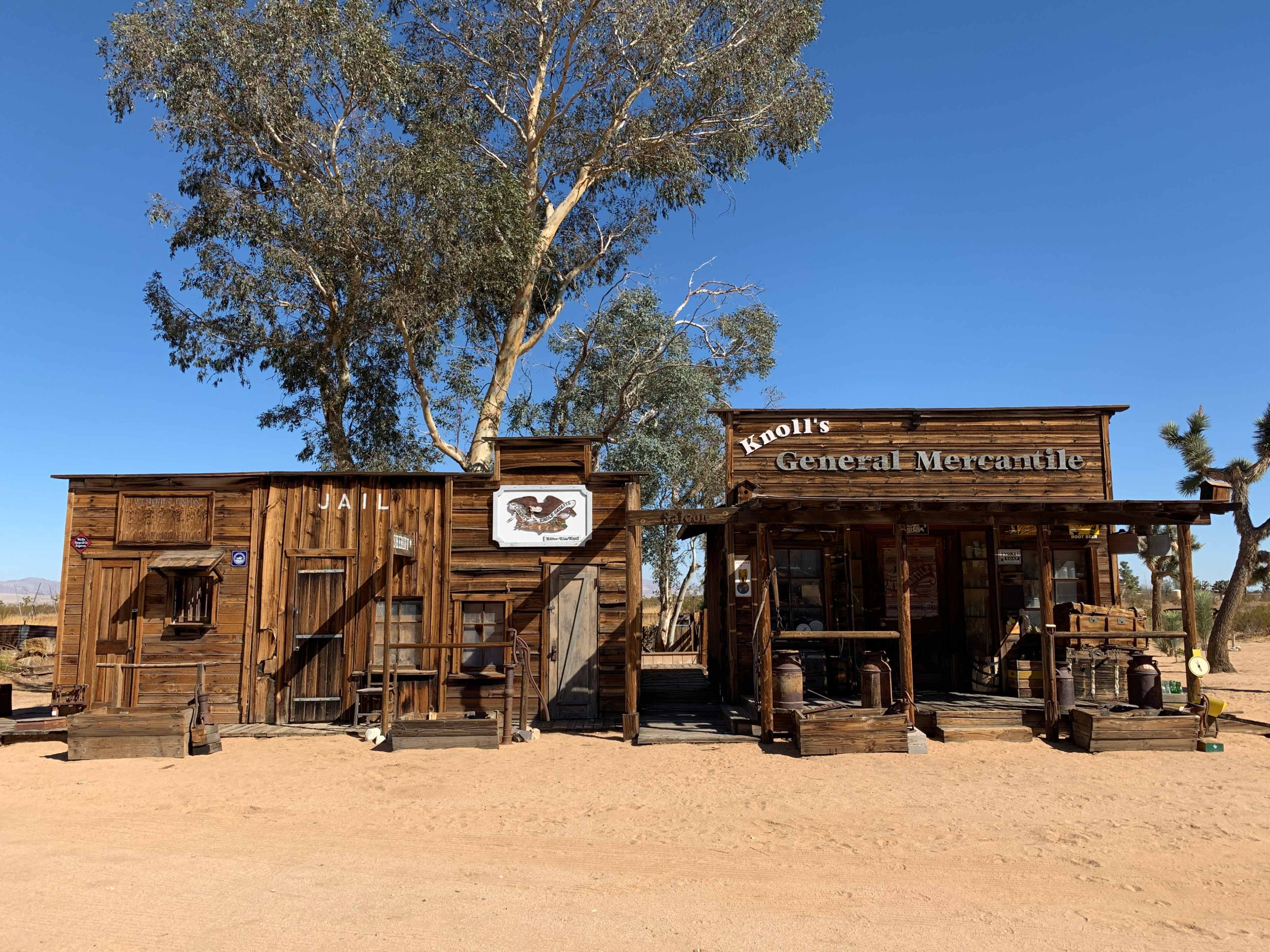 A rustic general mercantile store and jail, both made of weathered wood, set against a clear blue sky and sandy ground in a desert landscape.