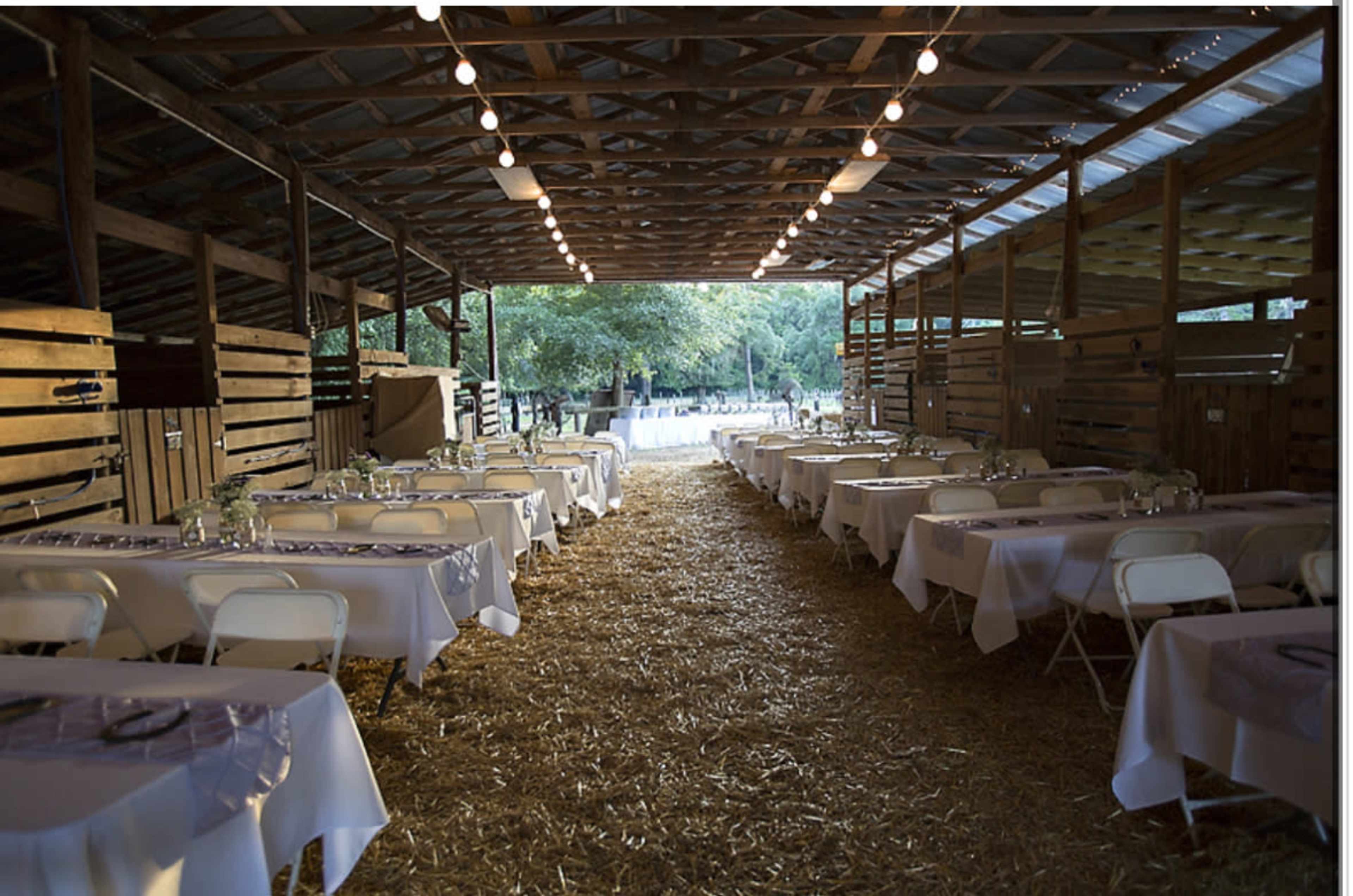 The image shows a rustic barn interior with long rows of tables set with white tablecloths and chairs, illuminated by strings of lights overhead.
