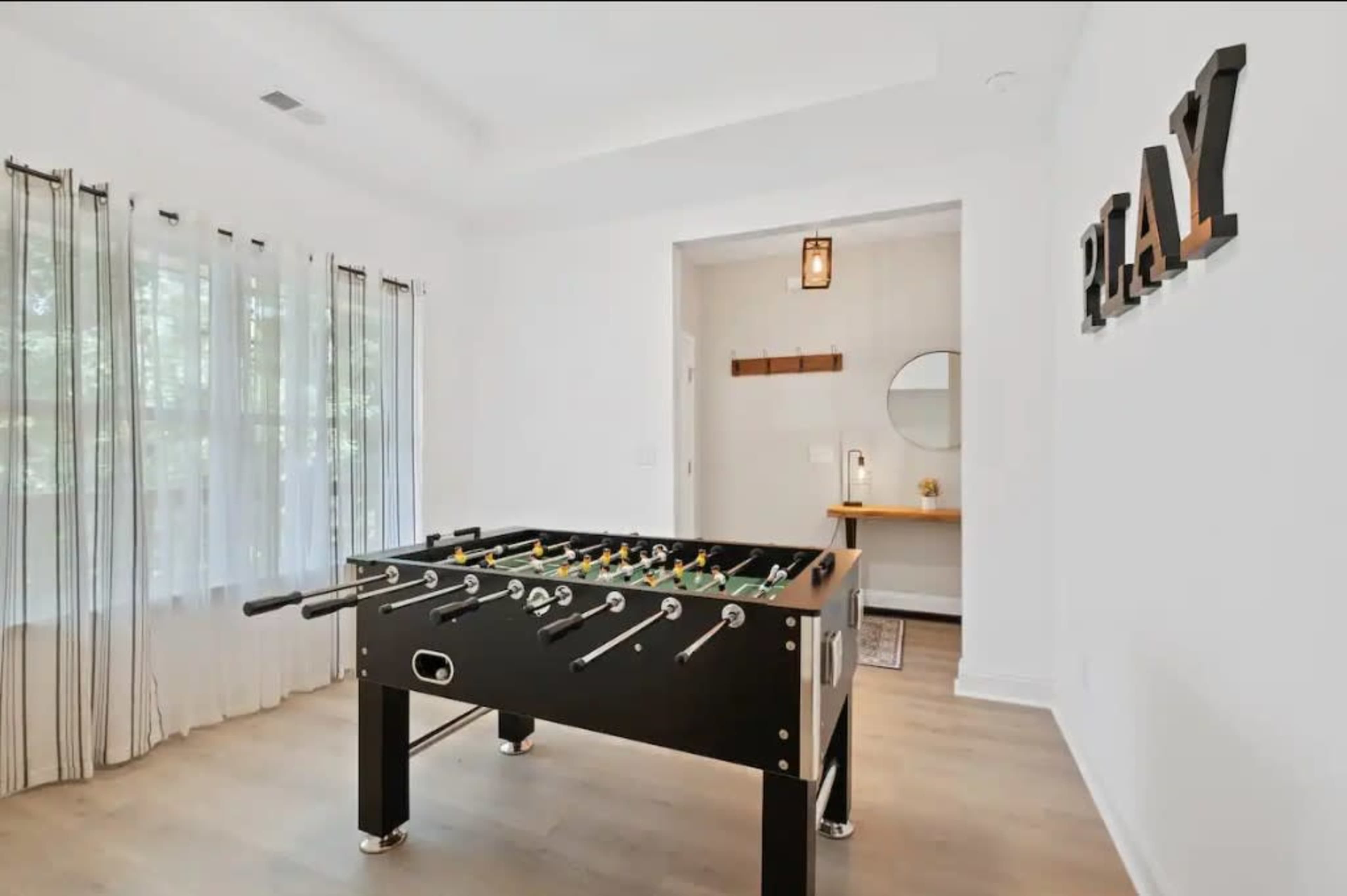 A foosball table is placed in a bright room with white walls and large windows, featuring a small table and mirror along one side.