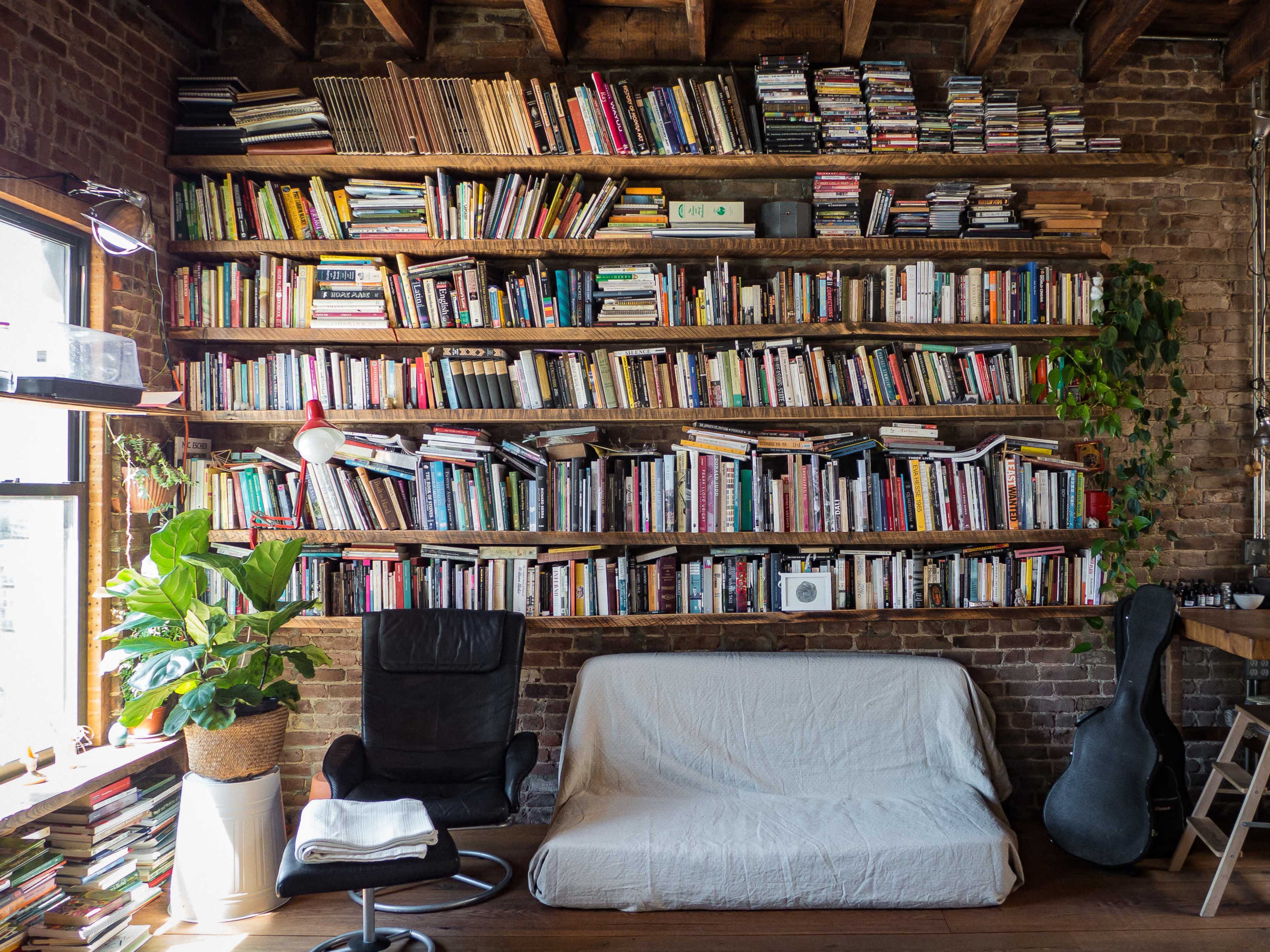 A cozy room features a wall of bookshelves filled with numerous books, a chair and a sofa covered with a white blanket, and a guitar leaning against the wall.