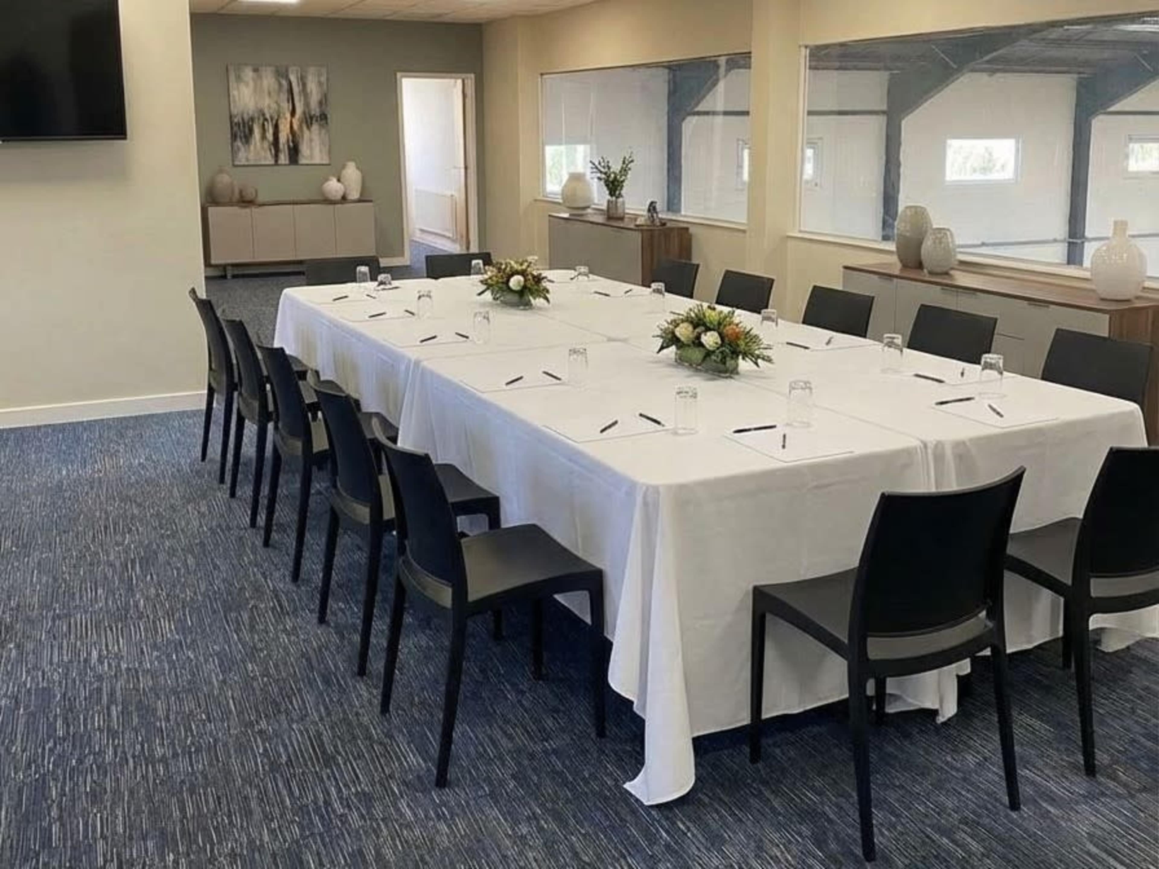 A large meeting room features a long table covered with a white tablecloth, surrounded by black chairs, and adorned with floral centerpieces.