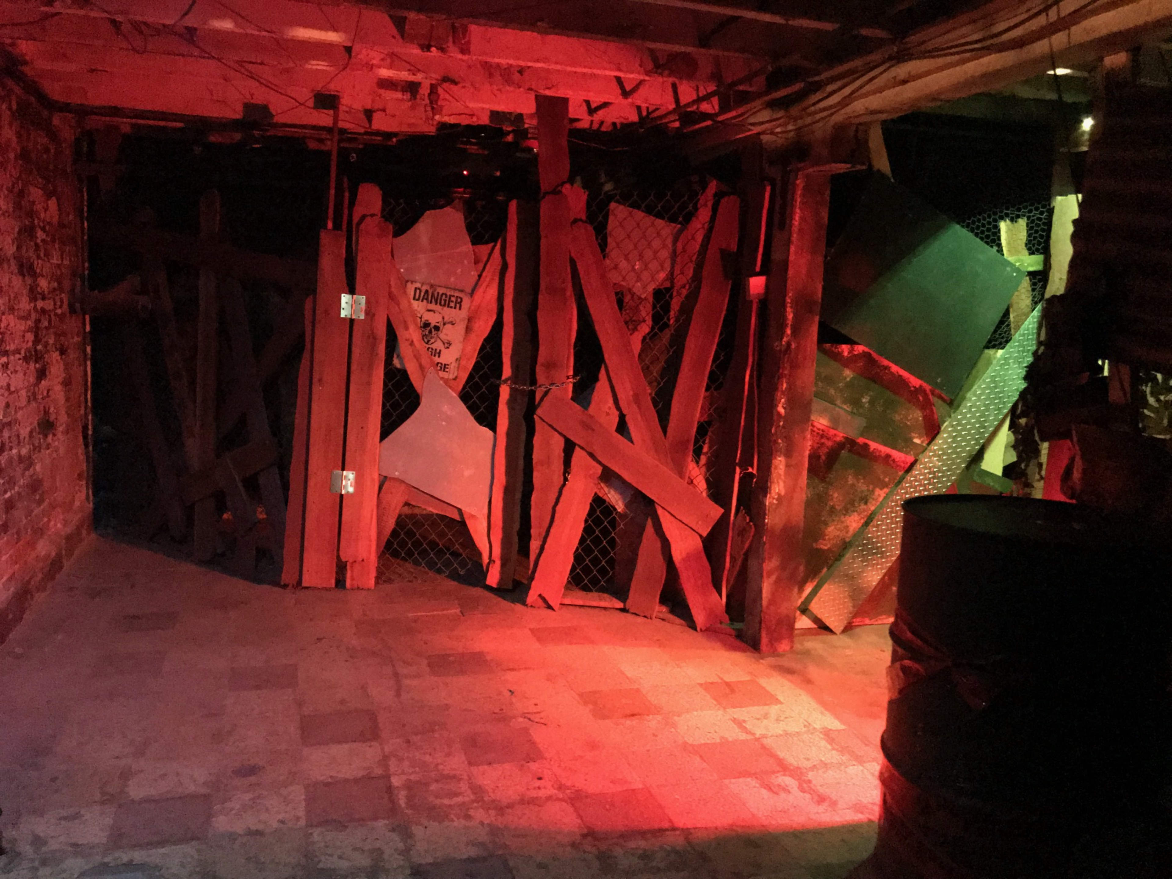 A dimly lit basement area with wooden barricades and a metal fence blocking a passage, illuminated by red lighting.
