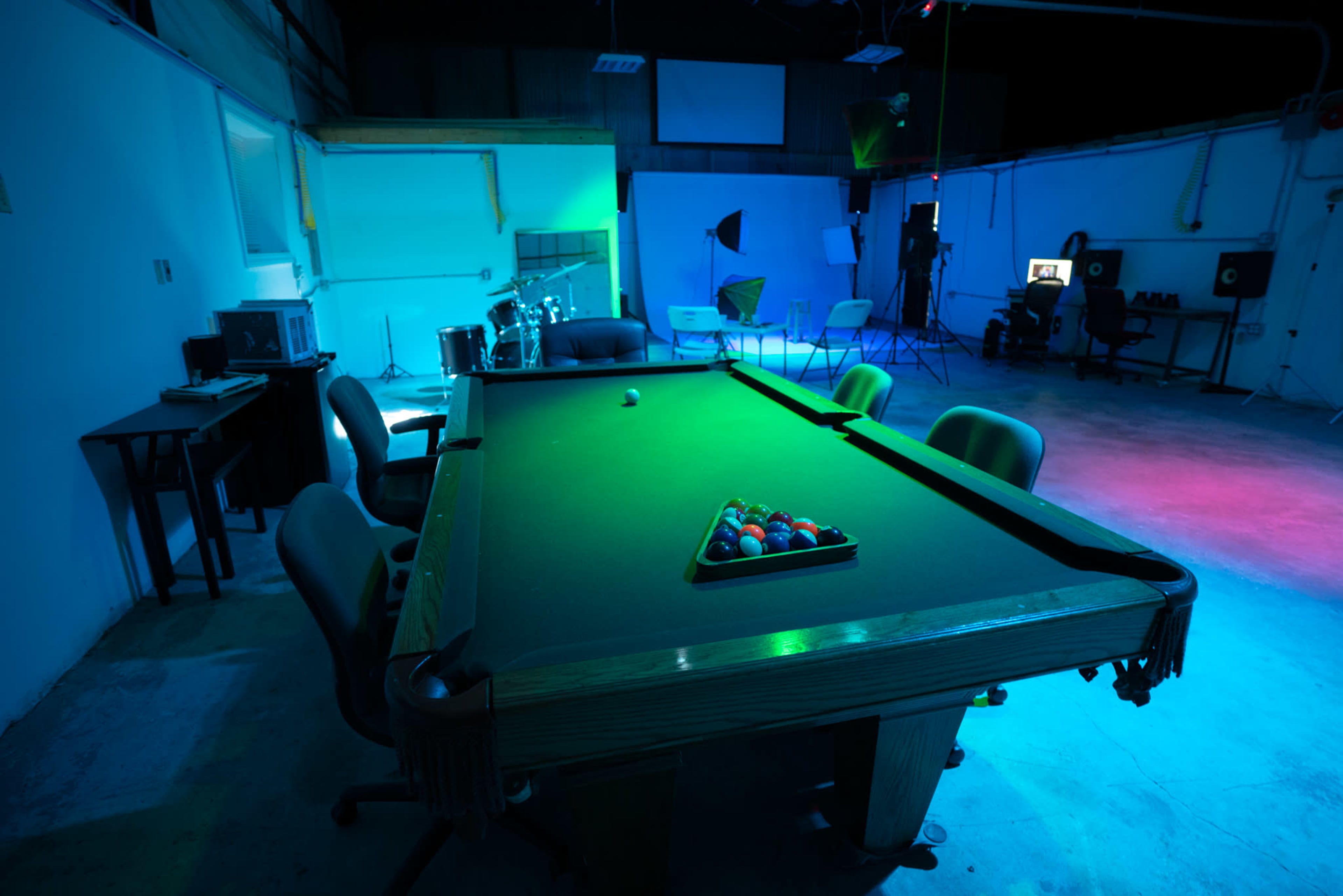 A billiard table with a full set of colored balls is positioned in a dimly lit room featuring studio equipment and seating areas.