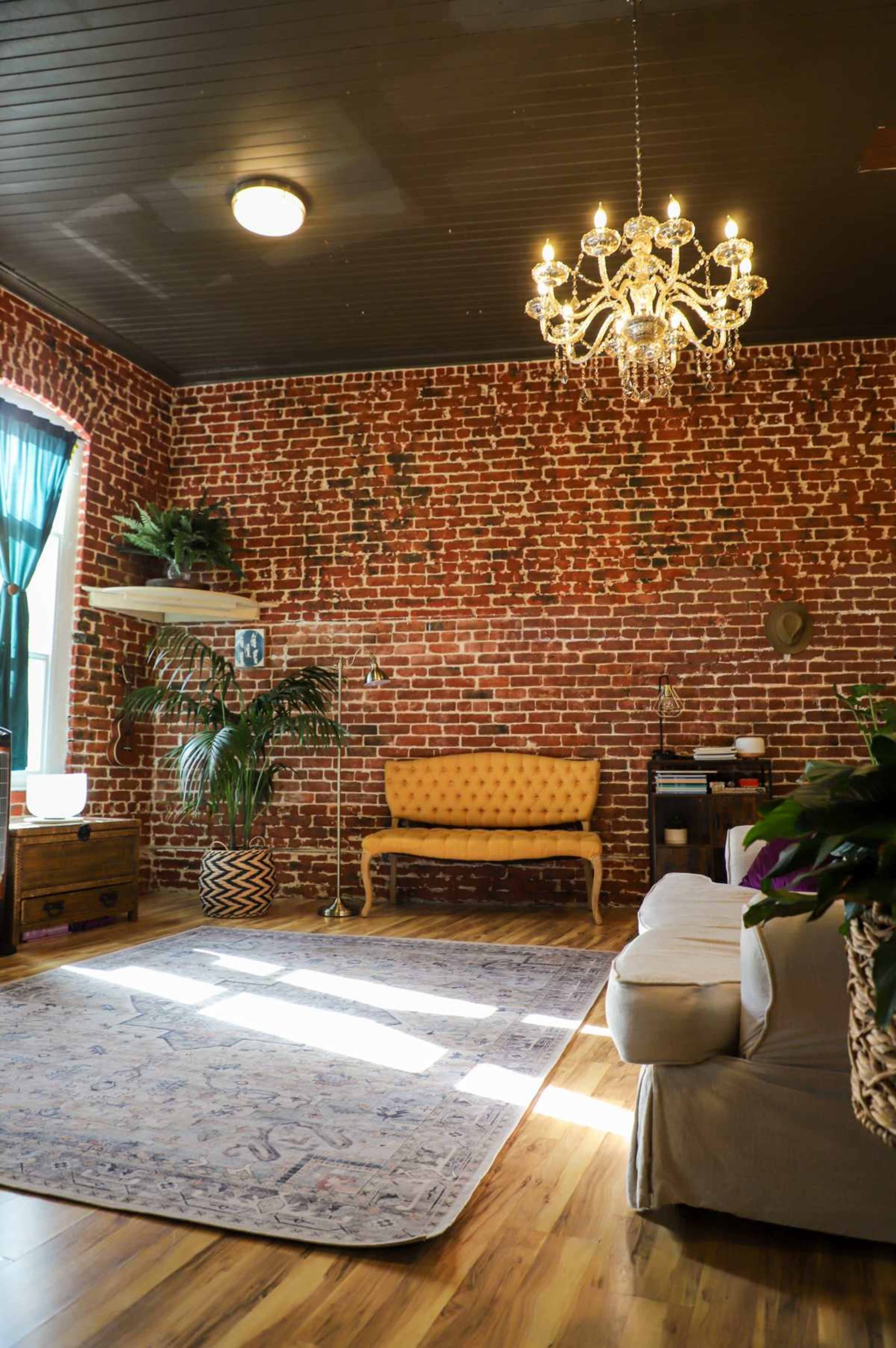 The image shows a spacious living room featuring exposed brick walls, a vintage chandelier, a yellow tufted sofa, and a decorative area rug on wooden flooring.