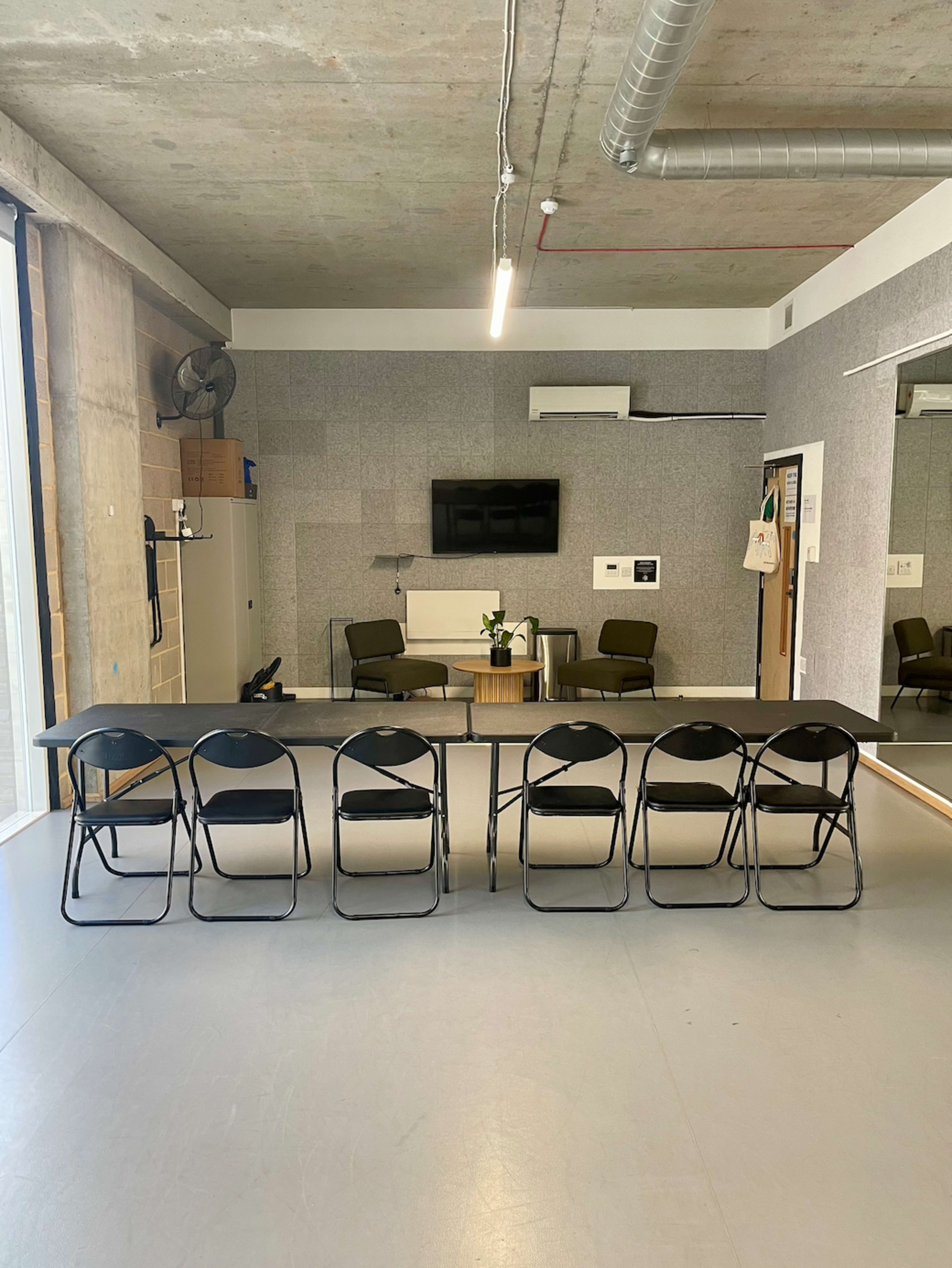 A spacious room with a long table and six black folding chairs arranged in a row, along with a small plant on the table and minimal furnishings in the background.