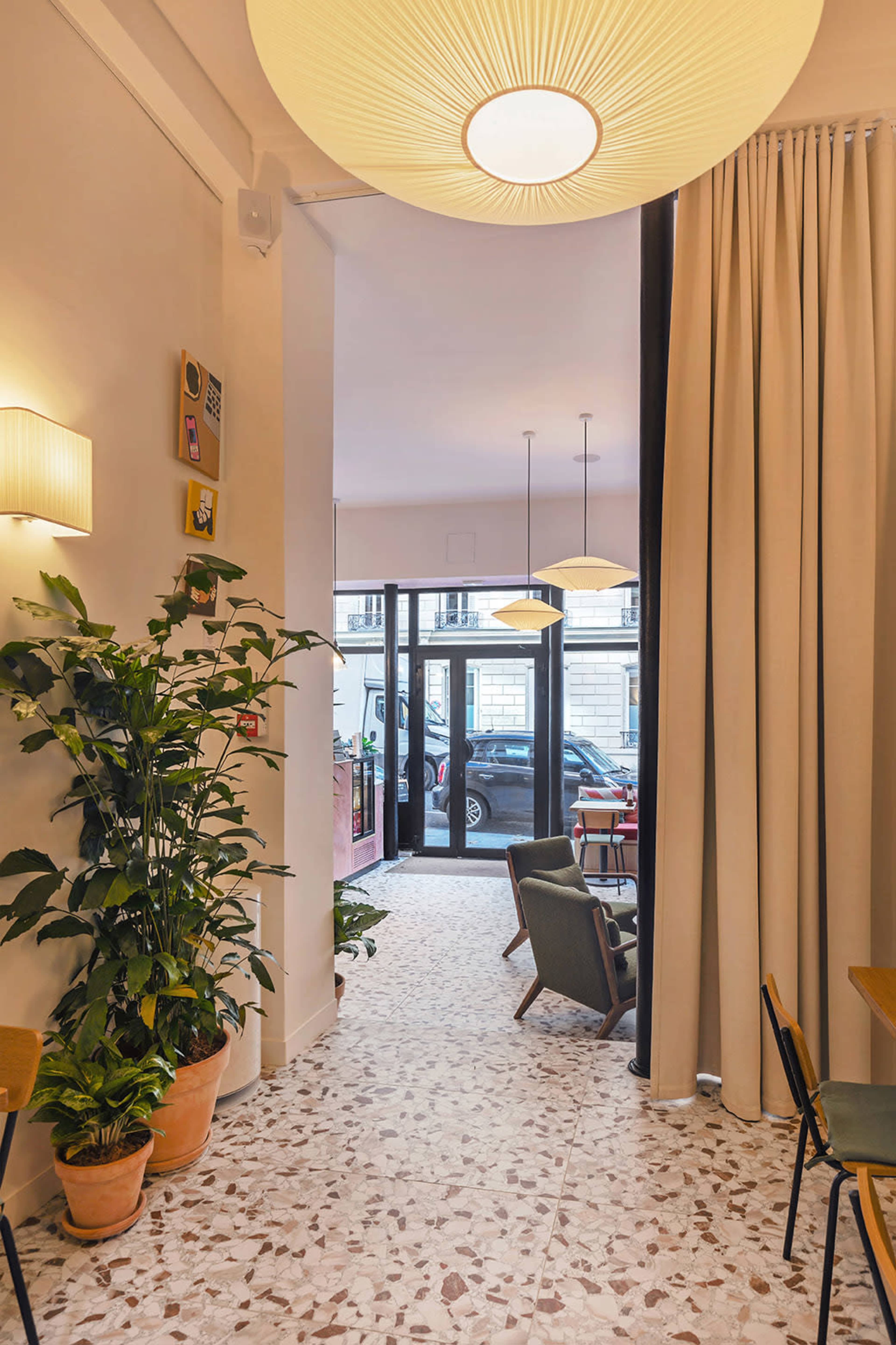 The image shows a modern café interior with a view of a seating area and a large window framed by curtains, complemented by decorative plants and pendant lighting.