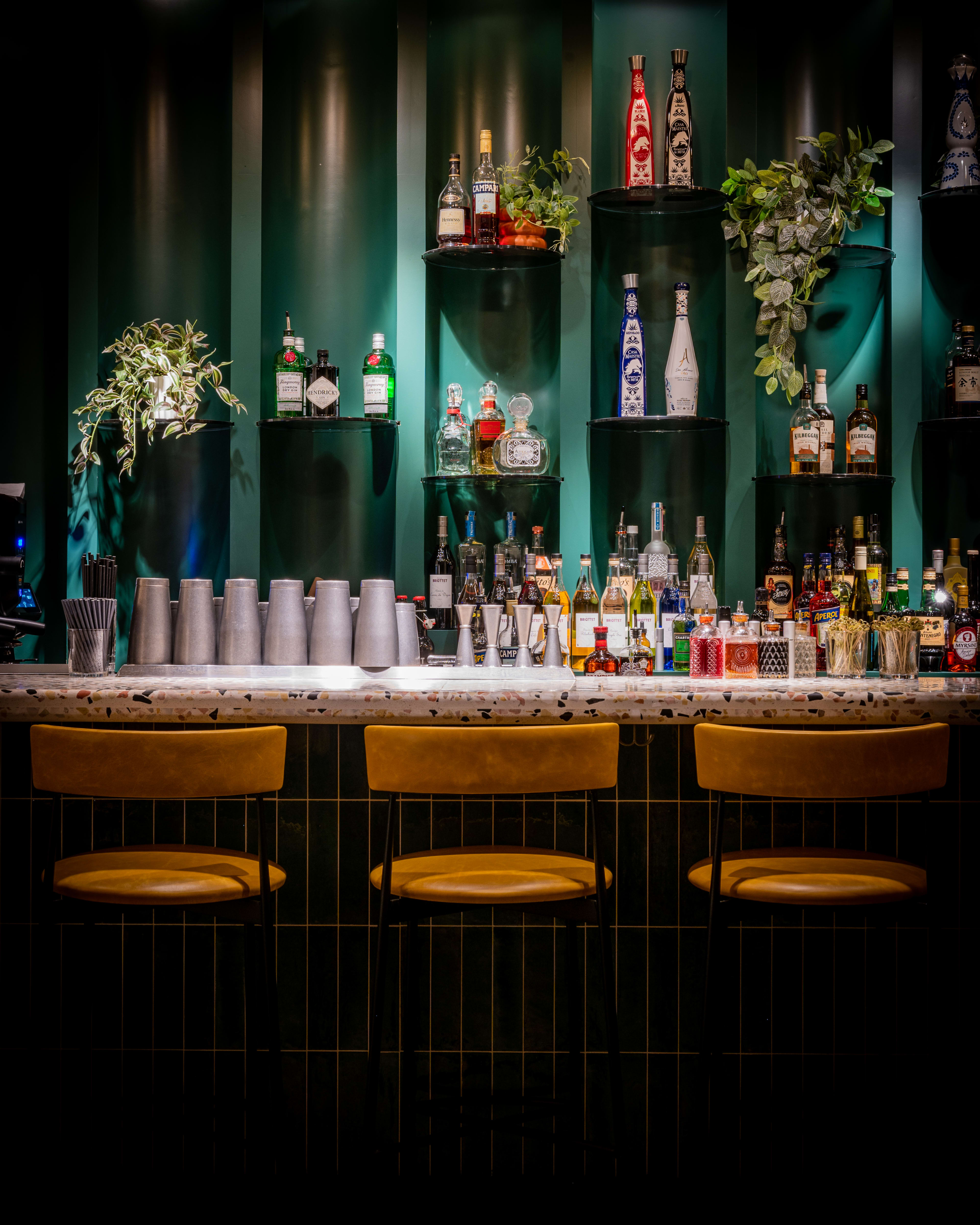 The image showcases a stylish bar with an array of illuminated bottles on shelves against a green wall, complemented by three empty chairs positioned at the counter.