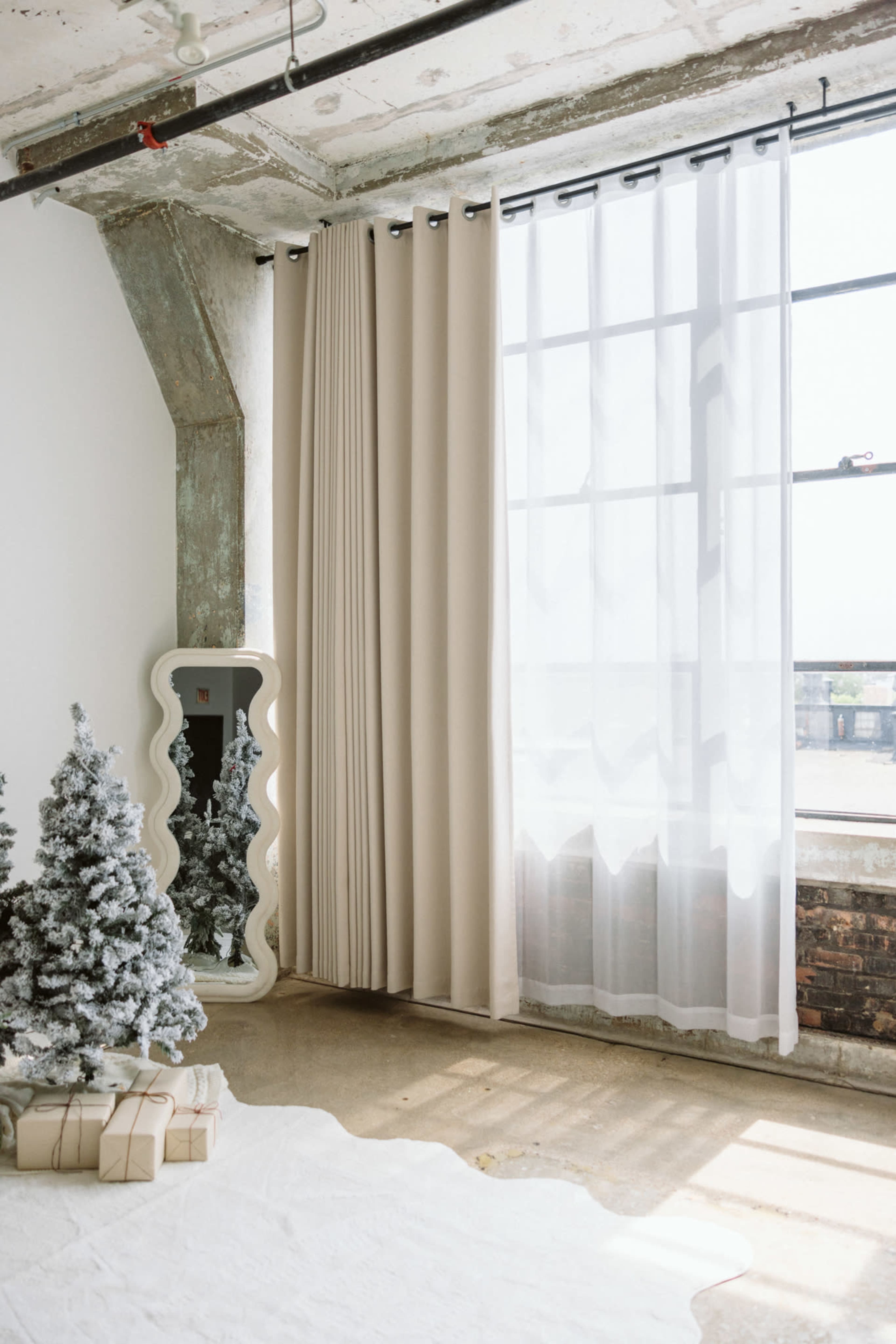 The image shows a well-lit room with large windows, featuring beige curtains, a decorative mirror, small artificial Christmas trees, and wrapped gifts on a white rug.