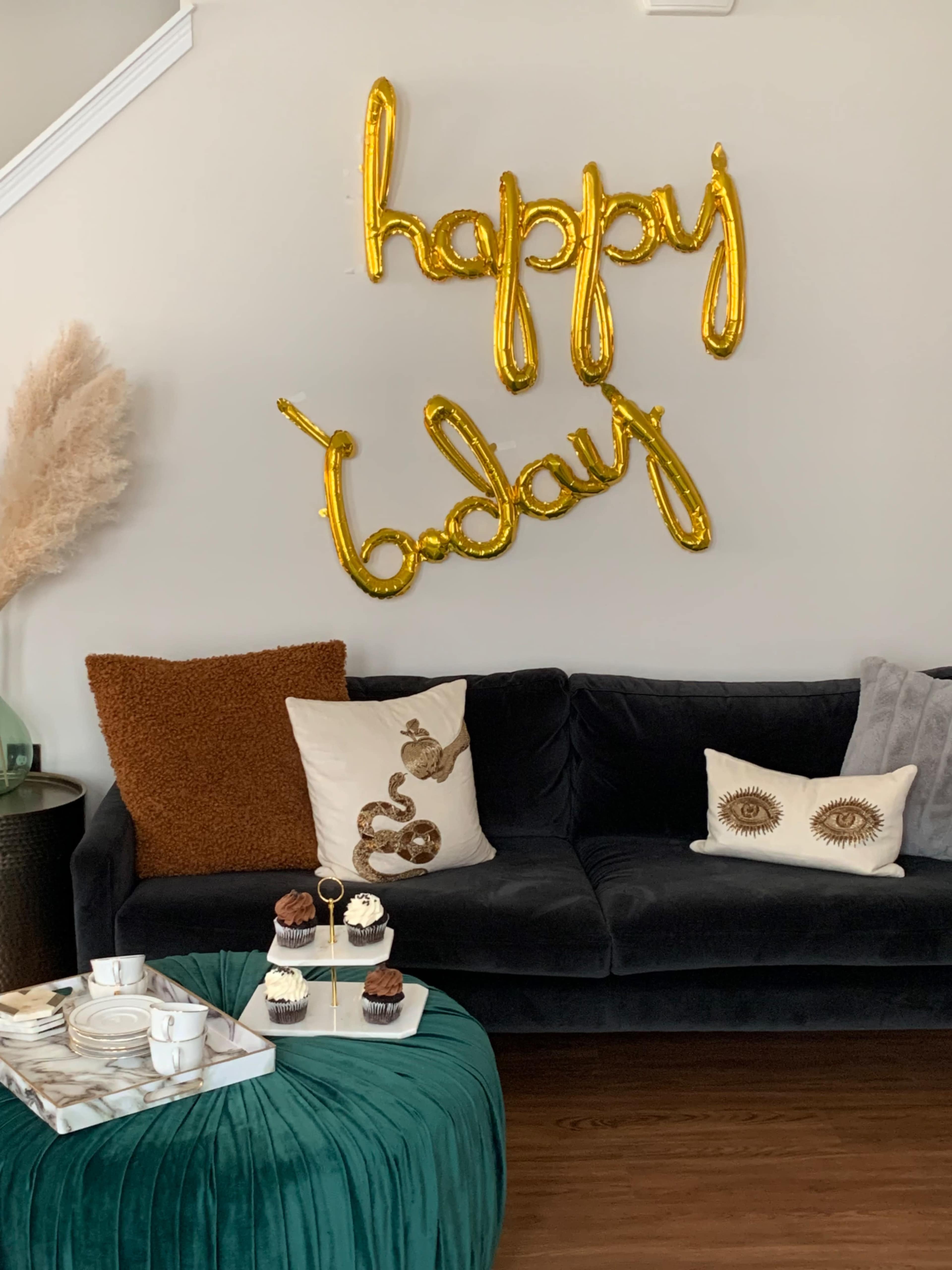 A cozy living room features a black sofa adorned with decorative pillows and a table displaying cupcakes, with golden balloon letters spelling "happy today" on the wall.
