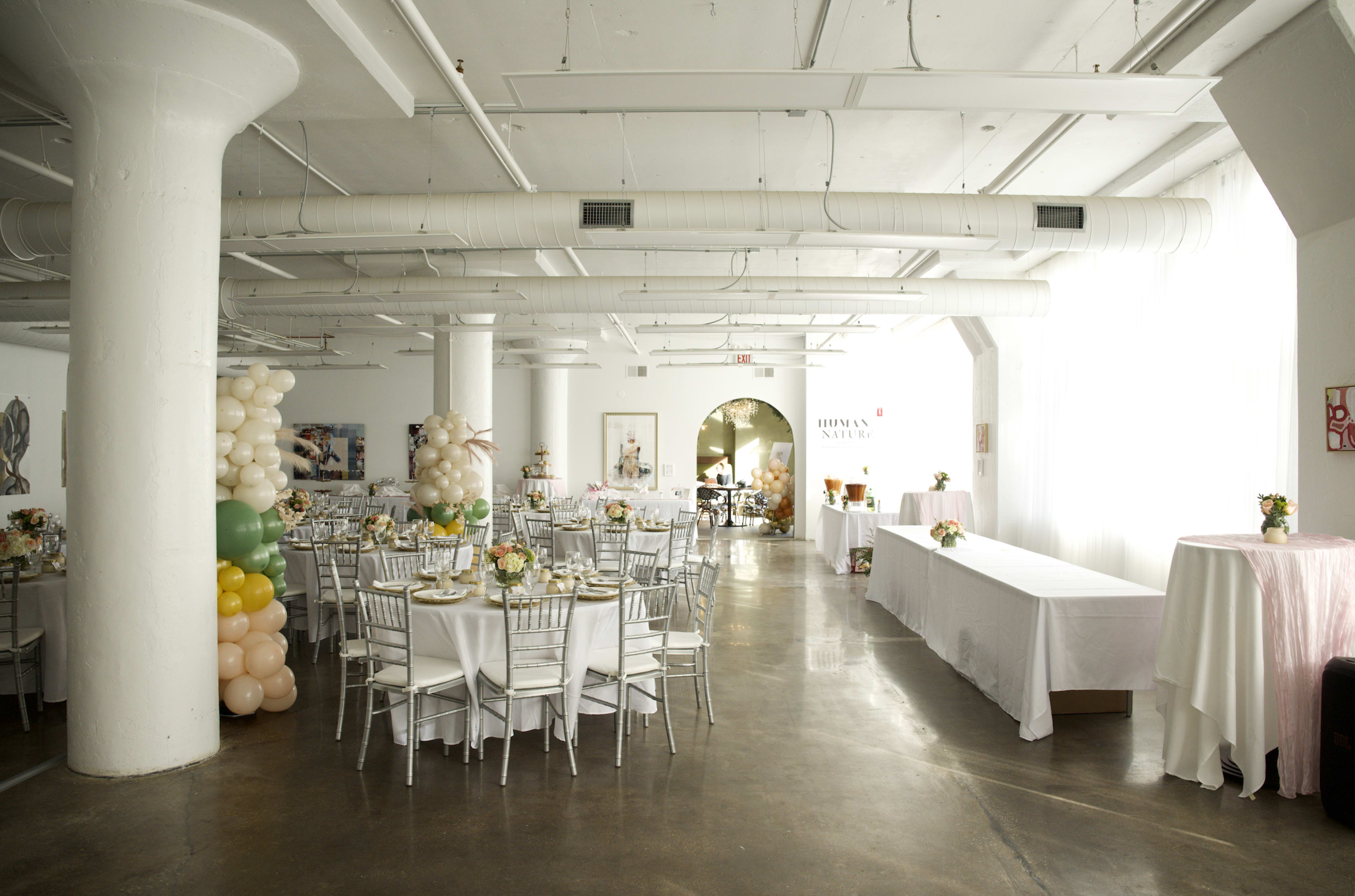The image shows a spacious event venue with round tables set for a gathering, decorated with flowers and balloons, and a serving table on one side.