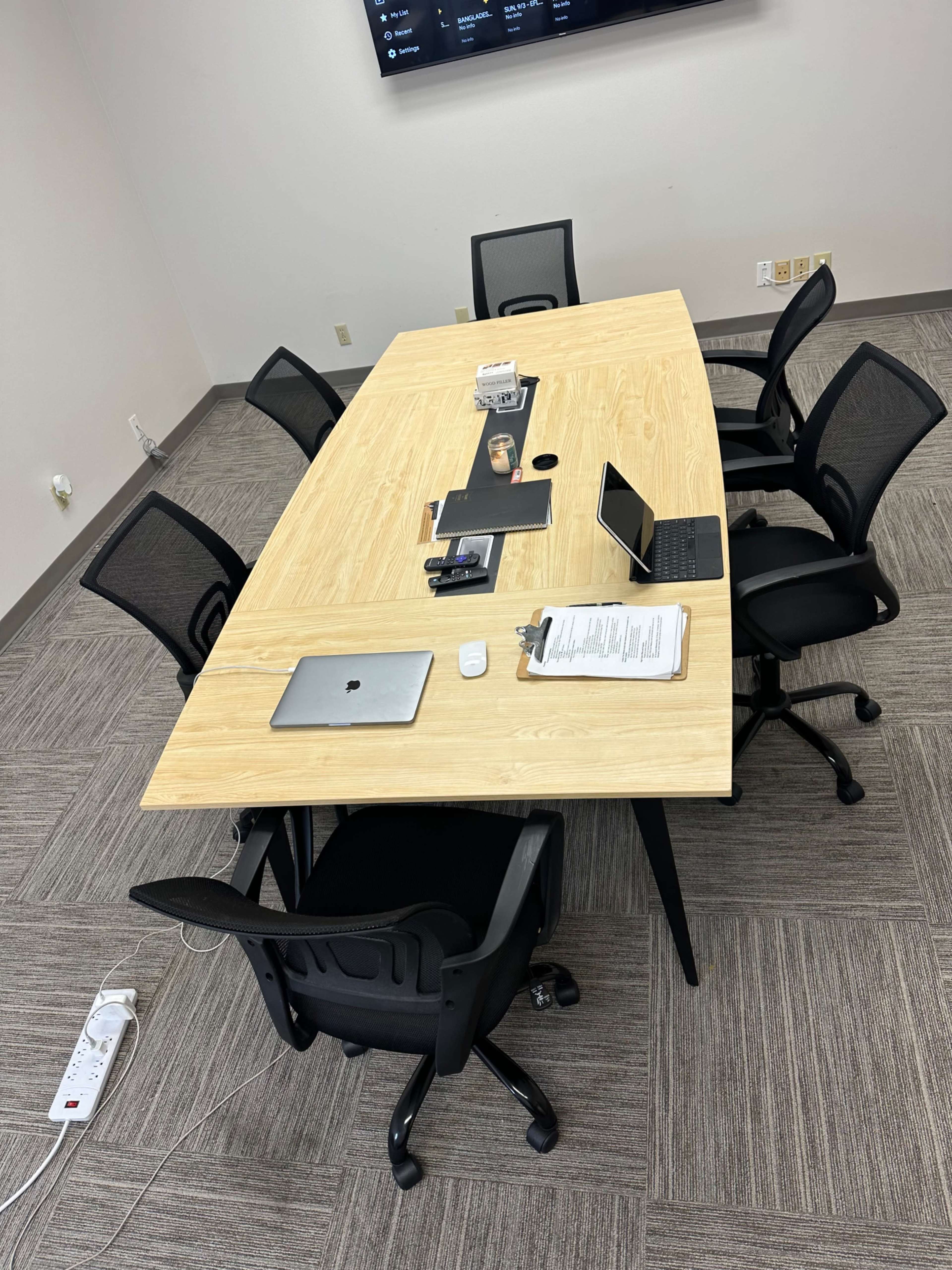 A large conference table with chairs surrounds it, and various office items including a laptop, tablet, and documents are placed on the table in a modern meeting room.