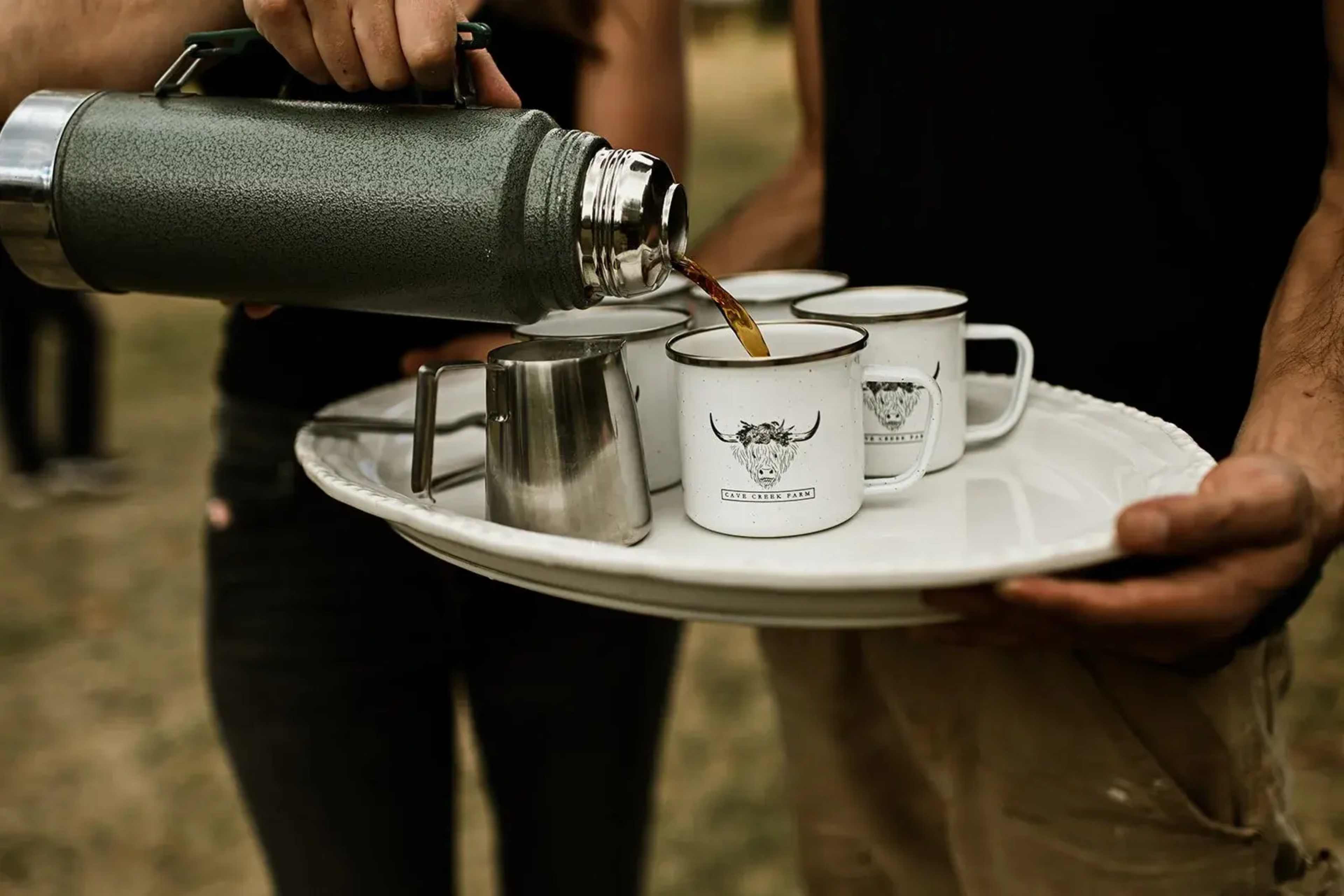 A person is pouring coffee from a thermos into several mugs arranged on a serving tray.