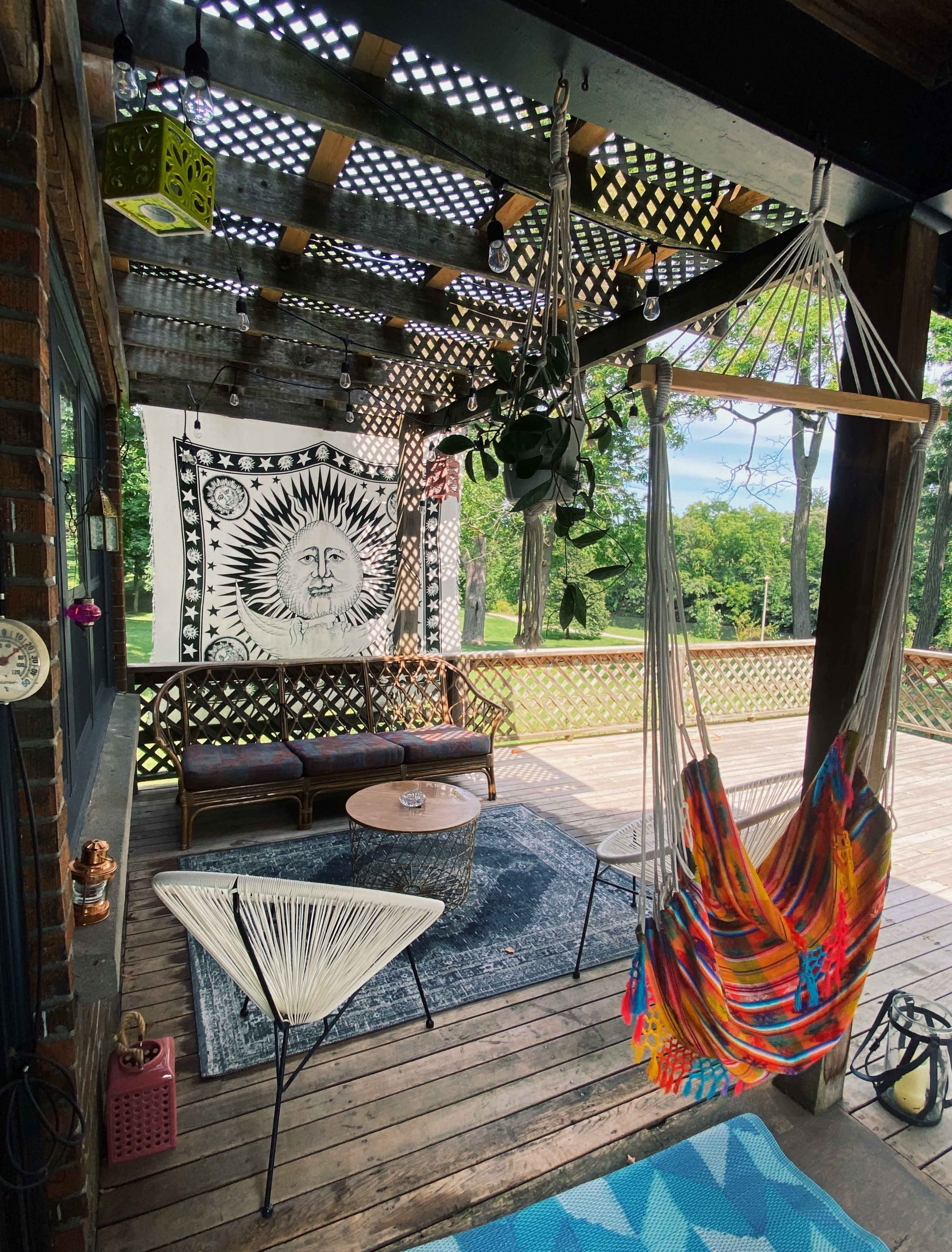 A covered porch with a sun-themed tapestry, colorful hammocks, and seating arrangements, surrounded by trees.
