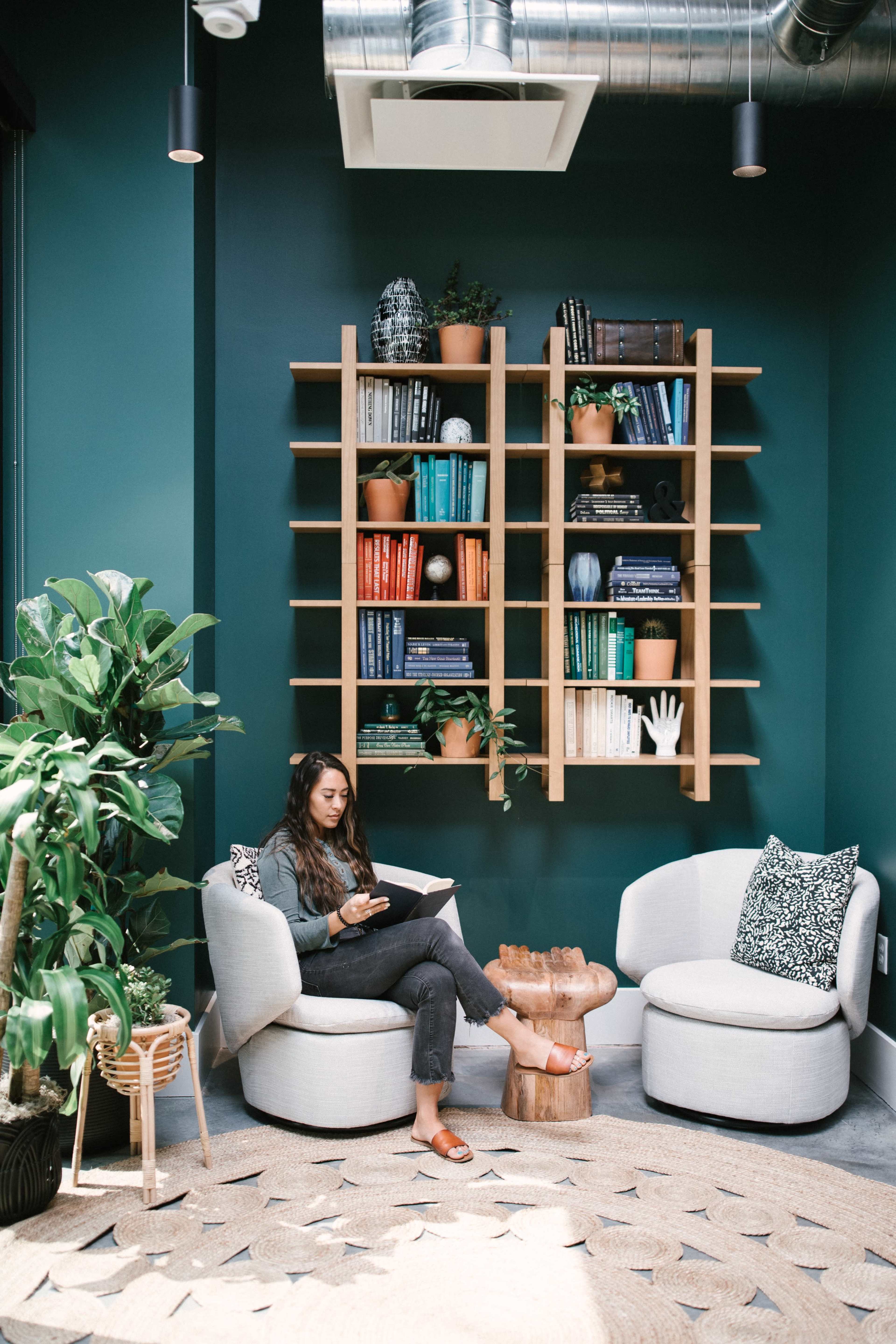 A woman sits in a modern, cozy reading area with a large bookshelf and potted plants around her.