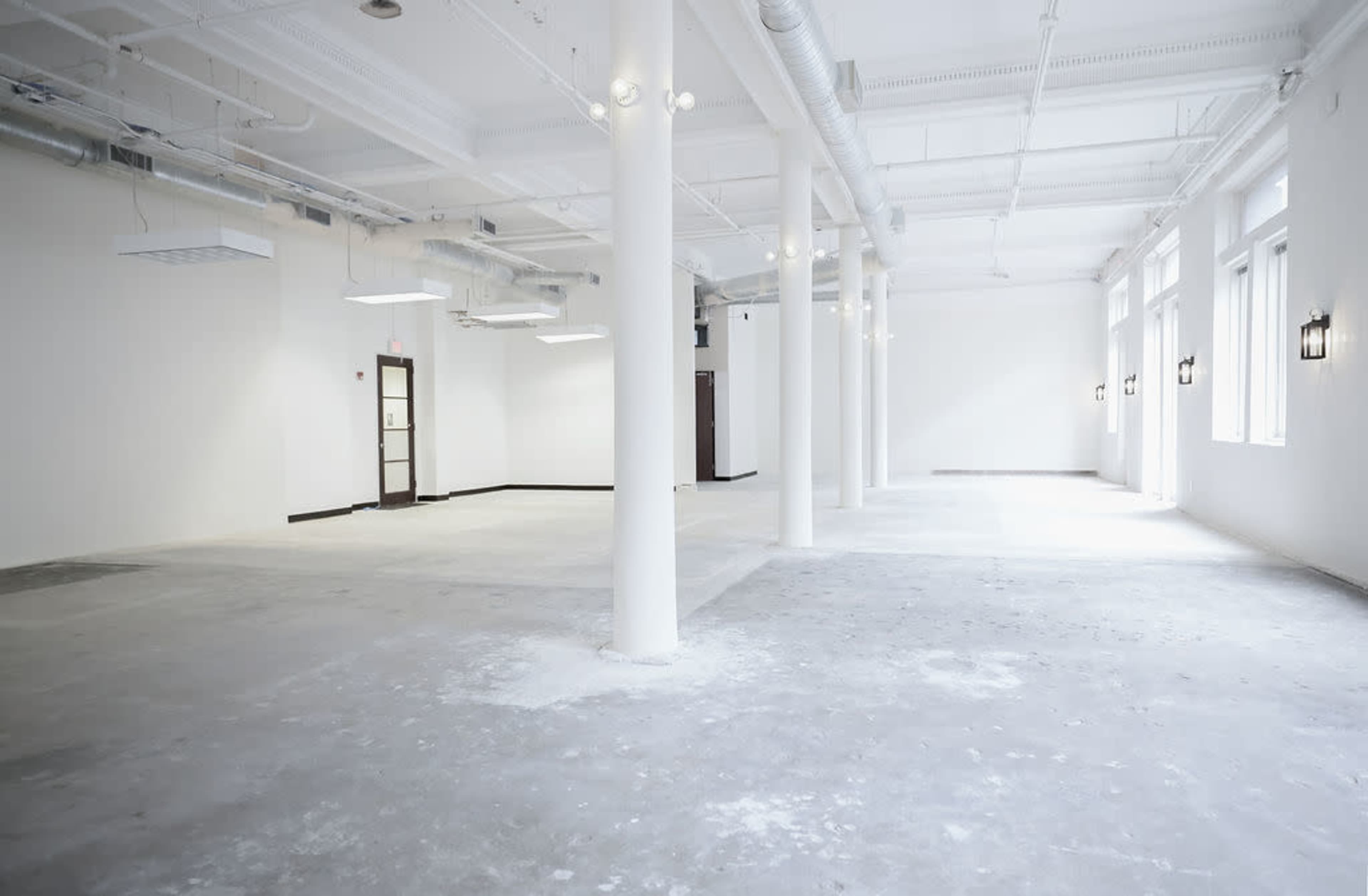 The image shows a spacious, empty room with white walls, exposed pipes, and large windows letting in natural light.