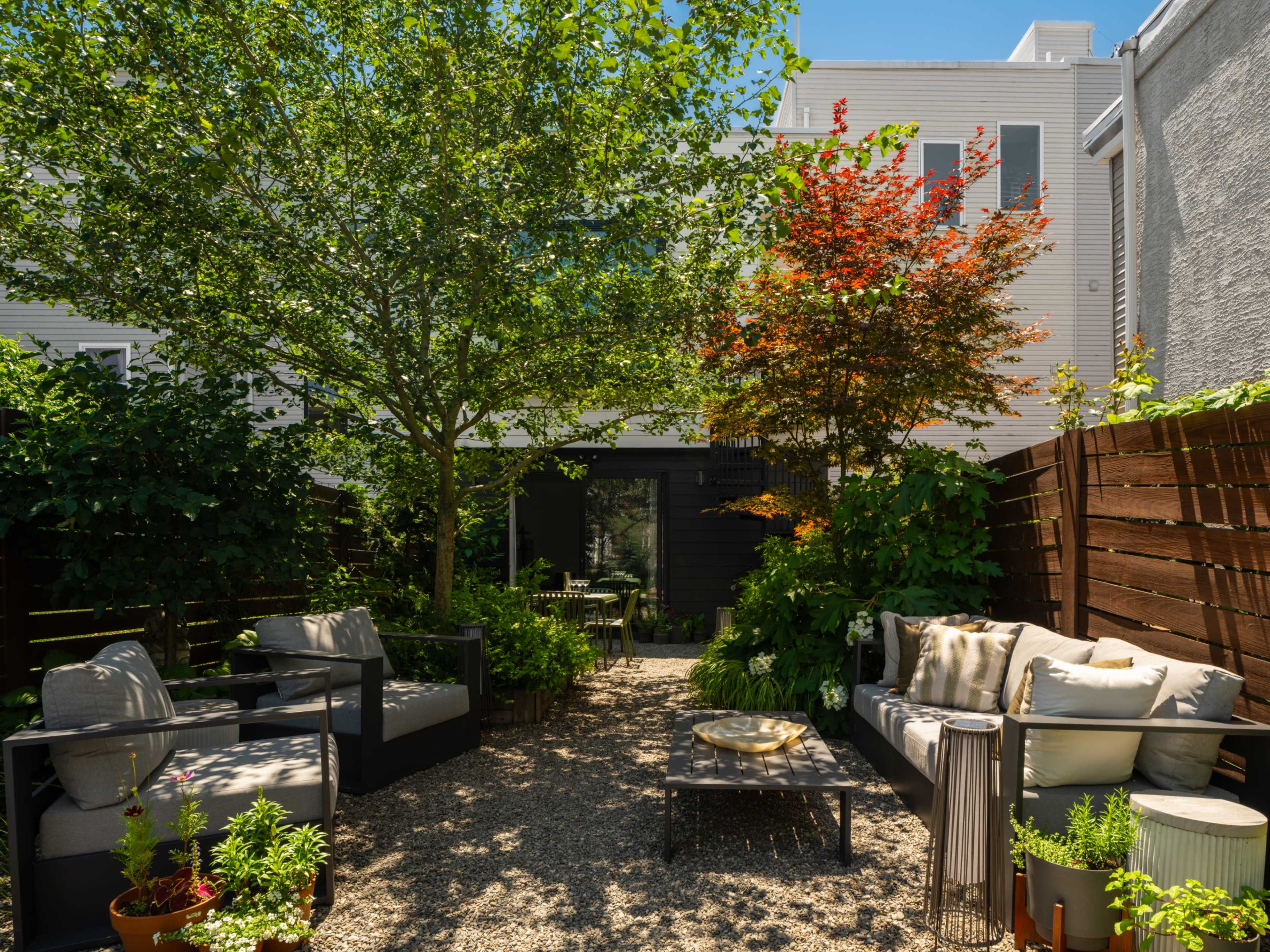 The image depicts a tranquil backyard garden featuring a seating area with sofas, a coffee table, and greenery, surrounded by wooden fences and a variety of plants.
