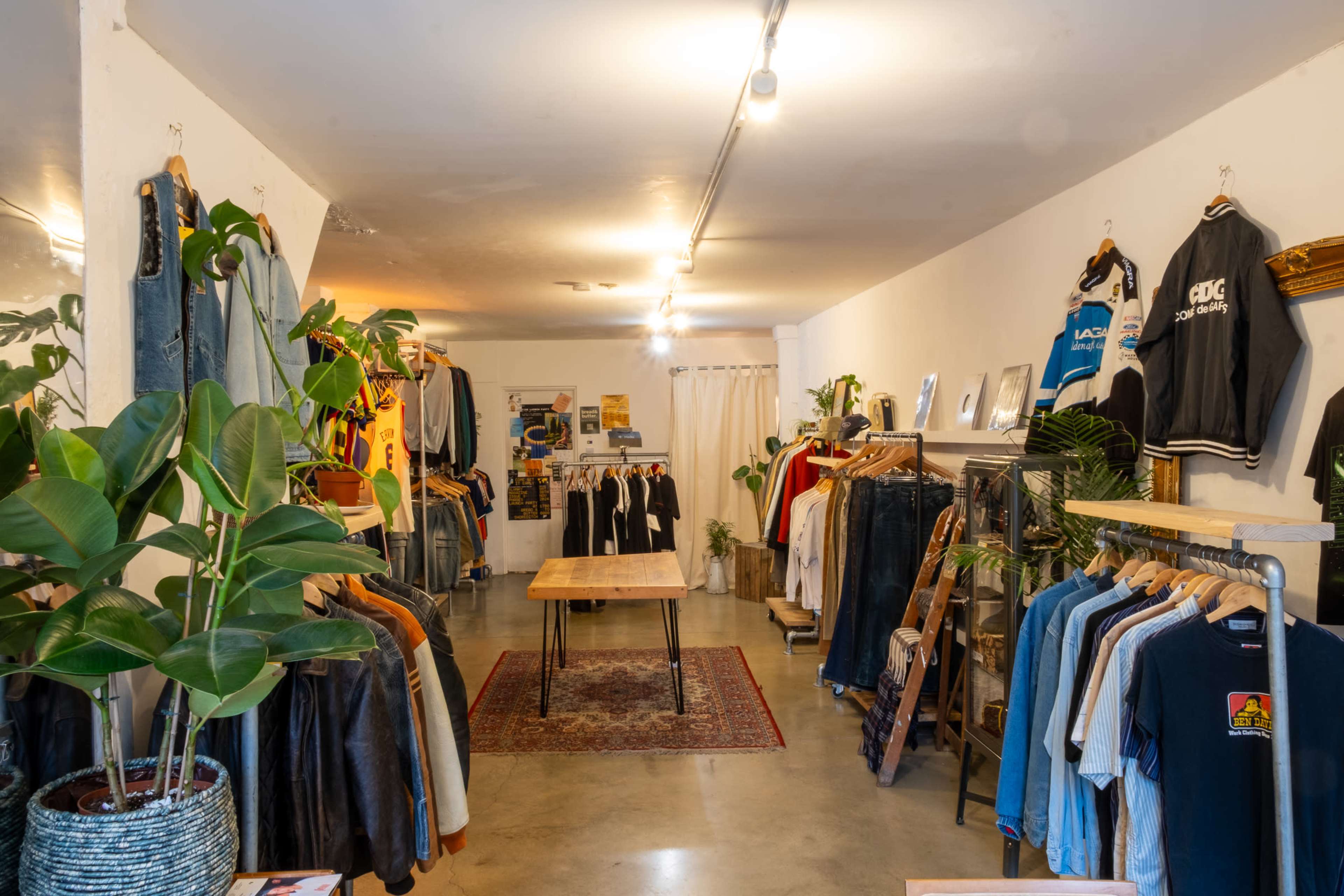 The image shows a clothing store interior featuring racks of various apparel, a wooden table in the center, and potted plants throughout.