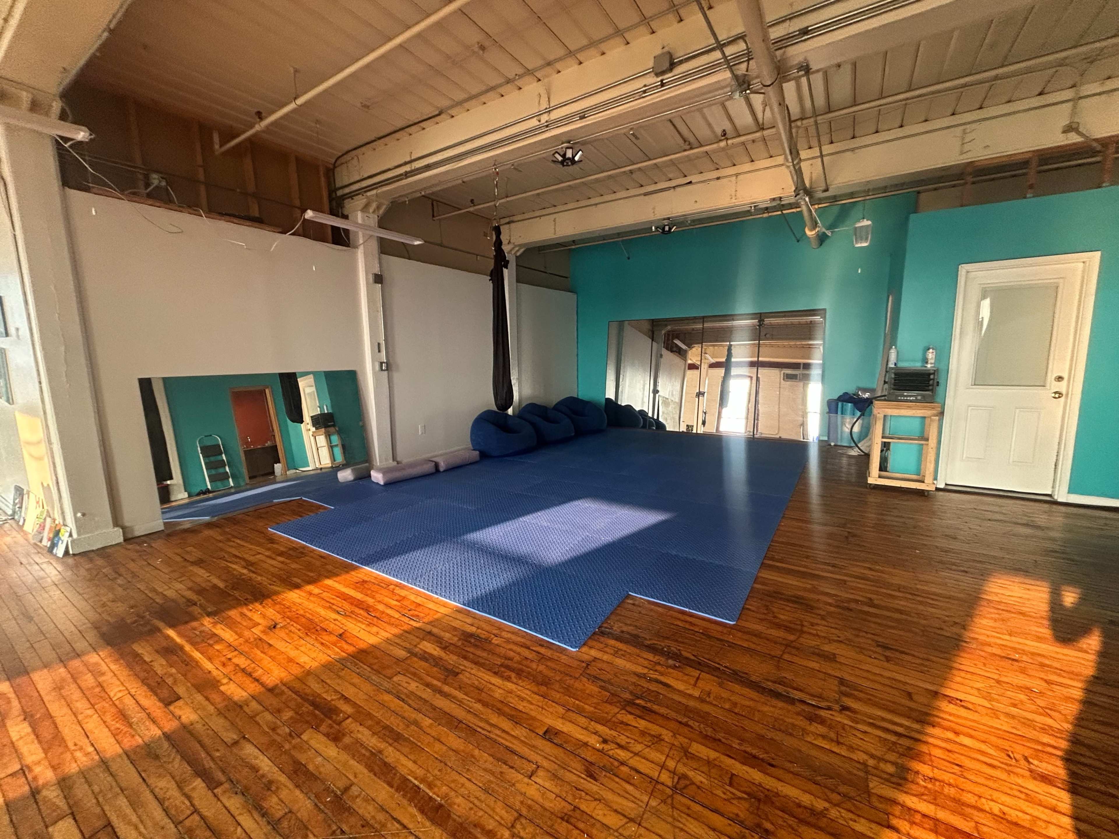 The image shows a spacious, empty yoga or exercise studio with blue mats on the floor and teal-painted walls.