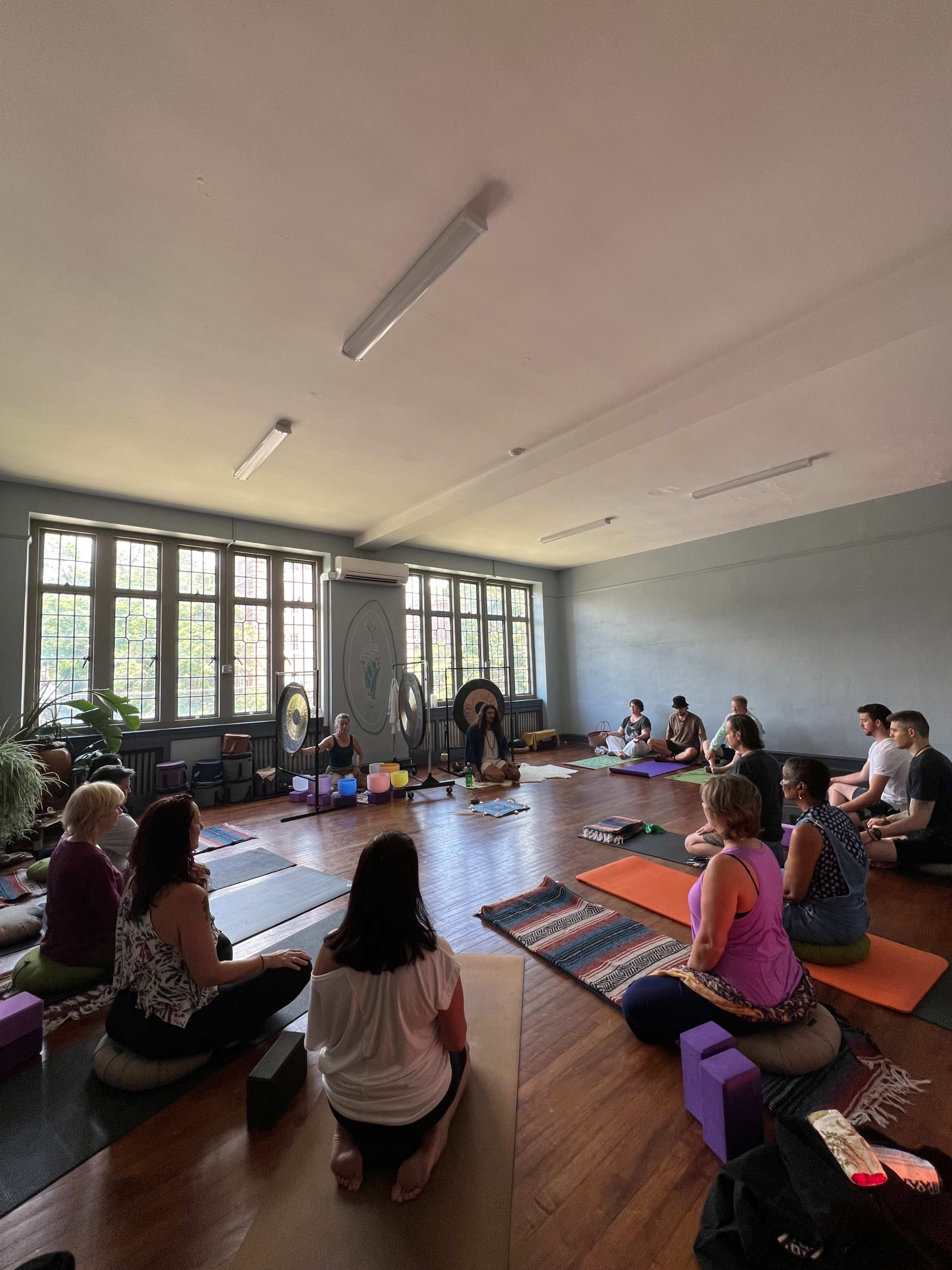 A group of people is seated on yoga mats and cushions in a spacious room with large windows, engaged in a yoga or meditation session.